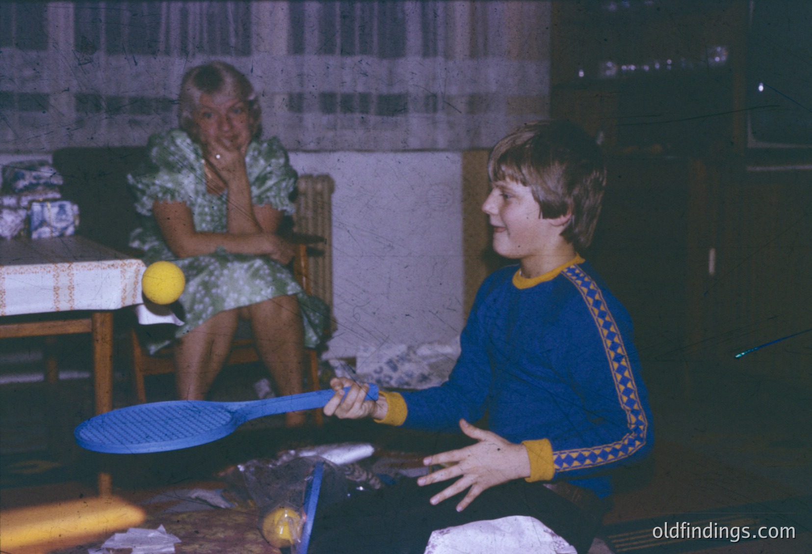 Vintage indoor ping-pong scene featuring two people in 1970s attire. Woman in floral dress observes while man in blue sweater with yellow trim prepares to strike a yellow ball with a paddle. Wooden table and basic furnishings suggest a home or community center setting.