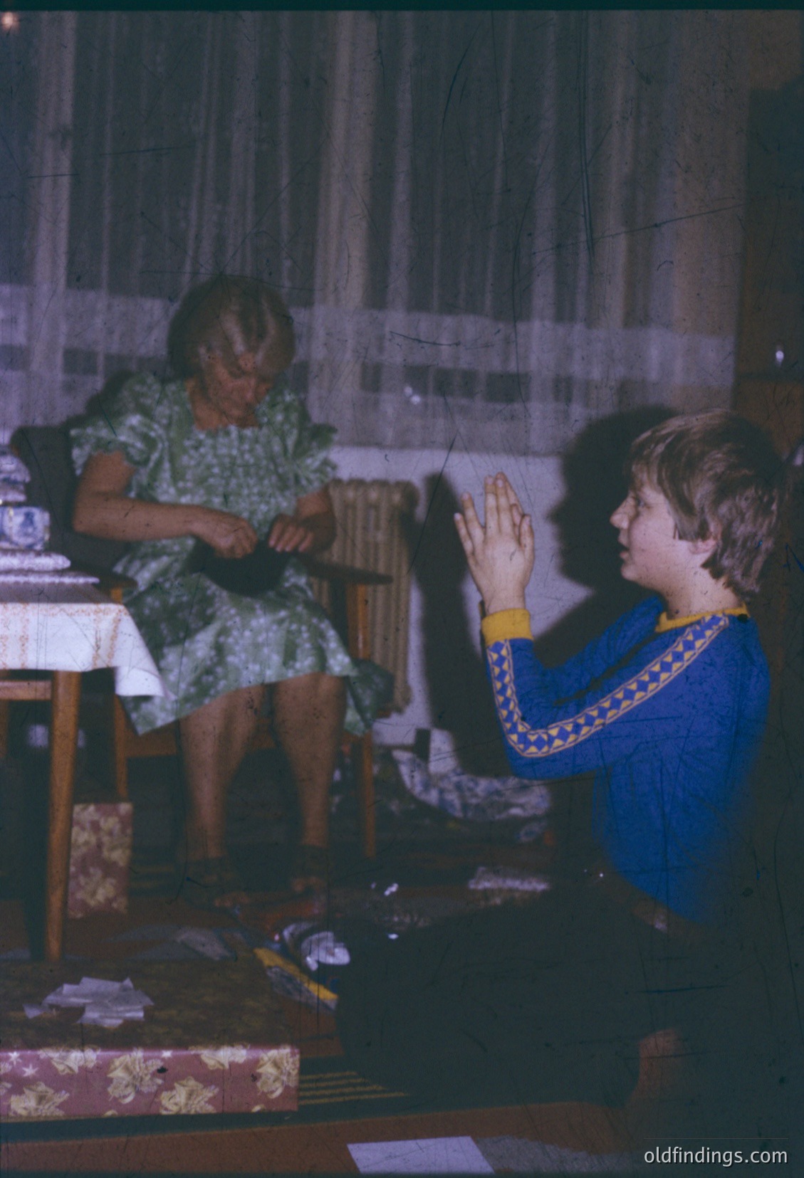 Vintage indoor scene featuring an elderly woman in floral dress sewing while a young boy in a blue embroidered shirt kneels beside her, hands clasped in prayer. Wooden furniture, patterned curtains, and a simple table setting suggest mid-20th century domestic life, likely Eastern European.