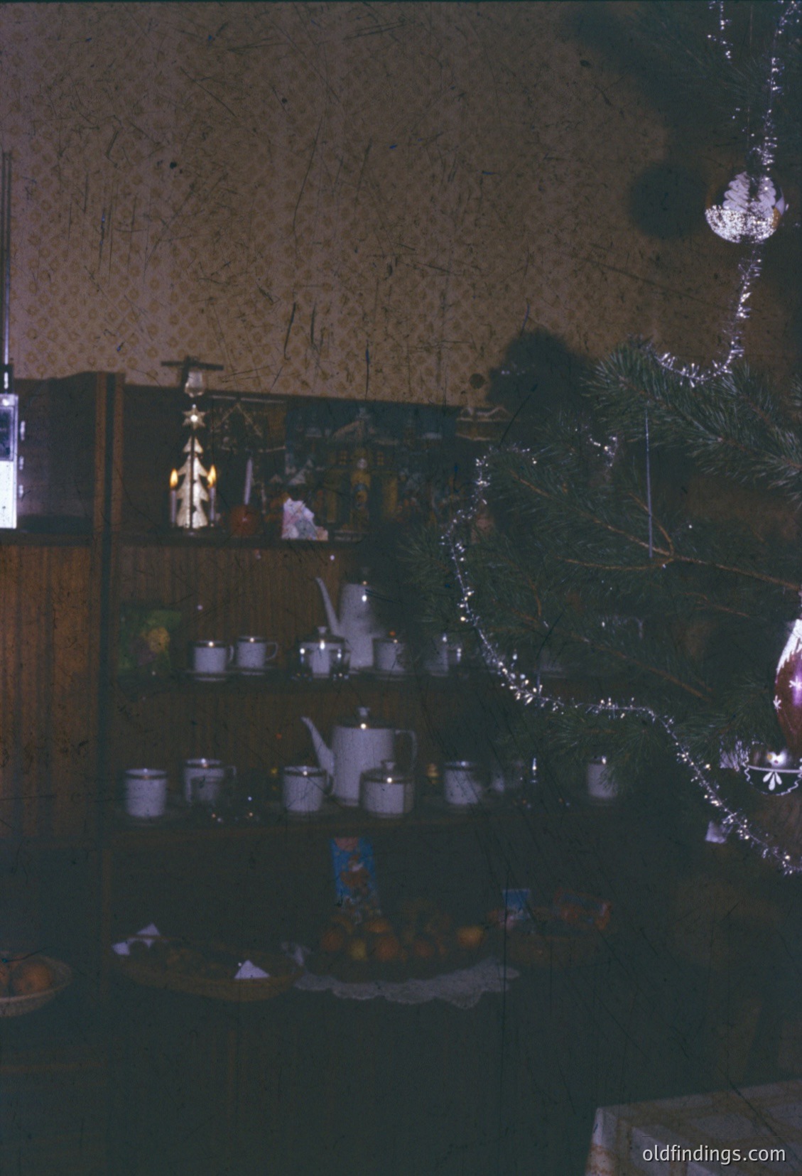Vintage holiday display featuring a wooden shelf adorned with silverware, ceramic teapots, and a tiered cake stand with festive ornaments. A small Christmas tree with lights and a star topper stands beside the shelf, illuminated by warm lighting. Mid-20th century European home decor, likely 1950s–1960s.