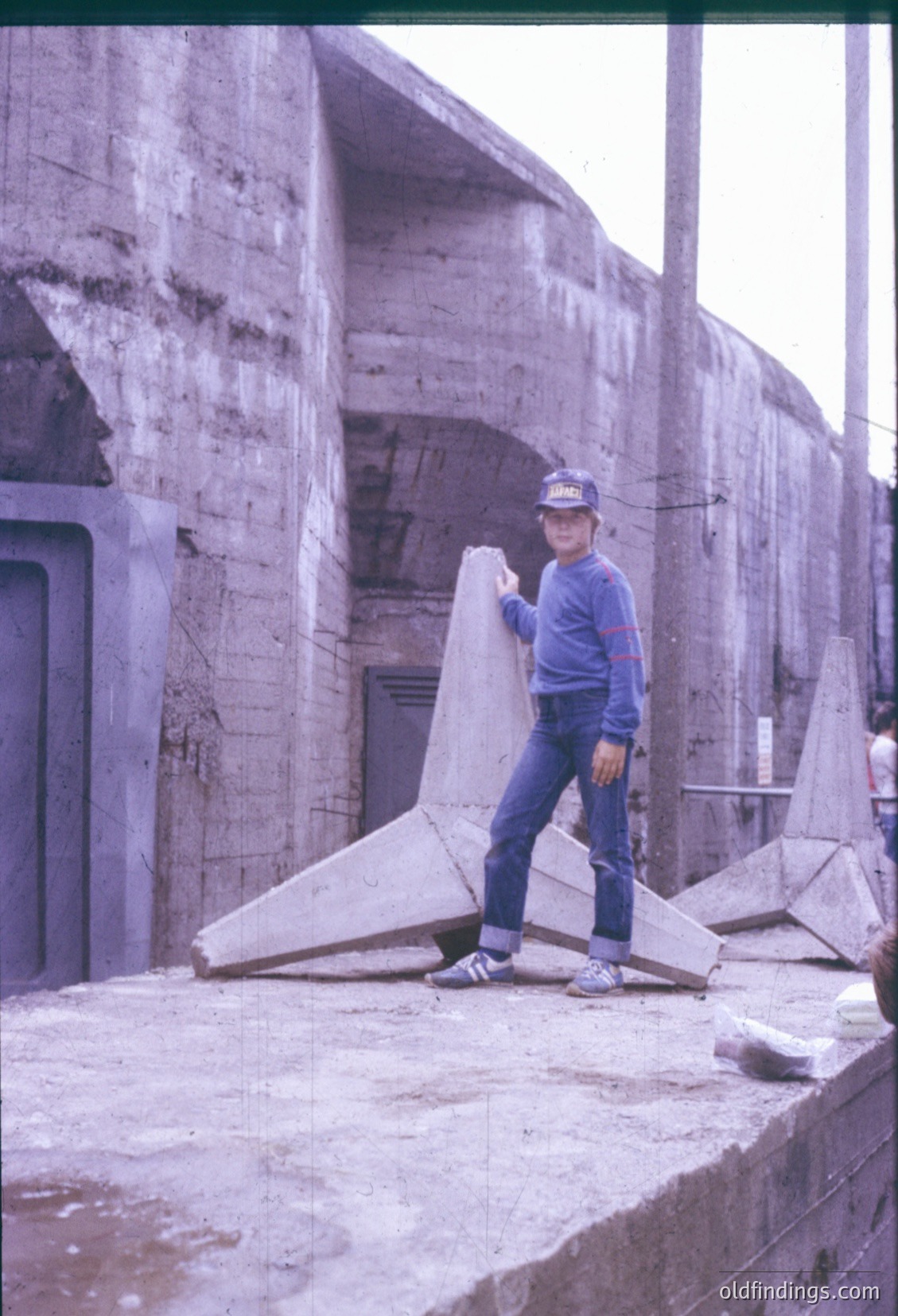 A man in 1970s-era casual wear (blue sweater, cap, jeans) poses beside a concrete structure resembling a coastal defense bunker or fortification. Broken concrete slabs lie scattered on a concrete platform, suggesting demolition or abandonment. The architecture hints at Cold War-era military infrastructure, likely Eastern Bloc.