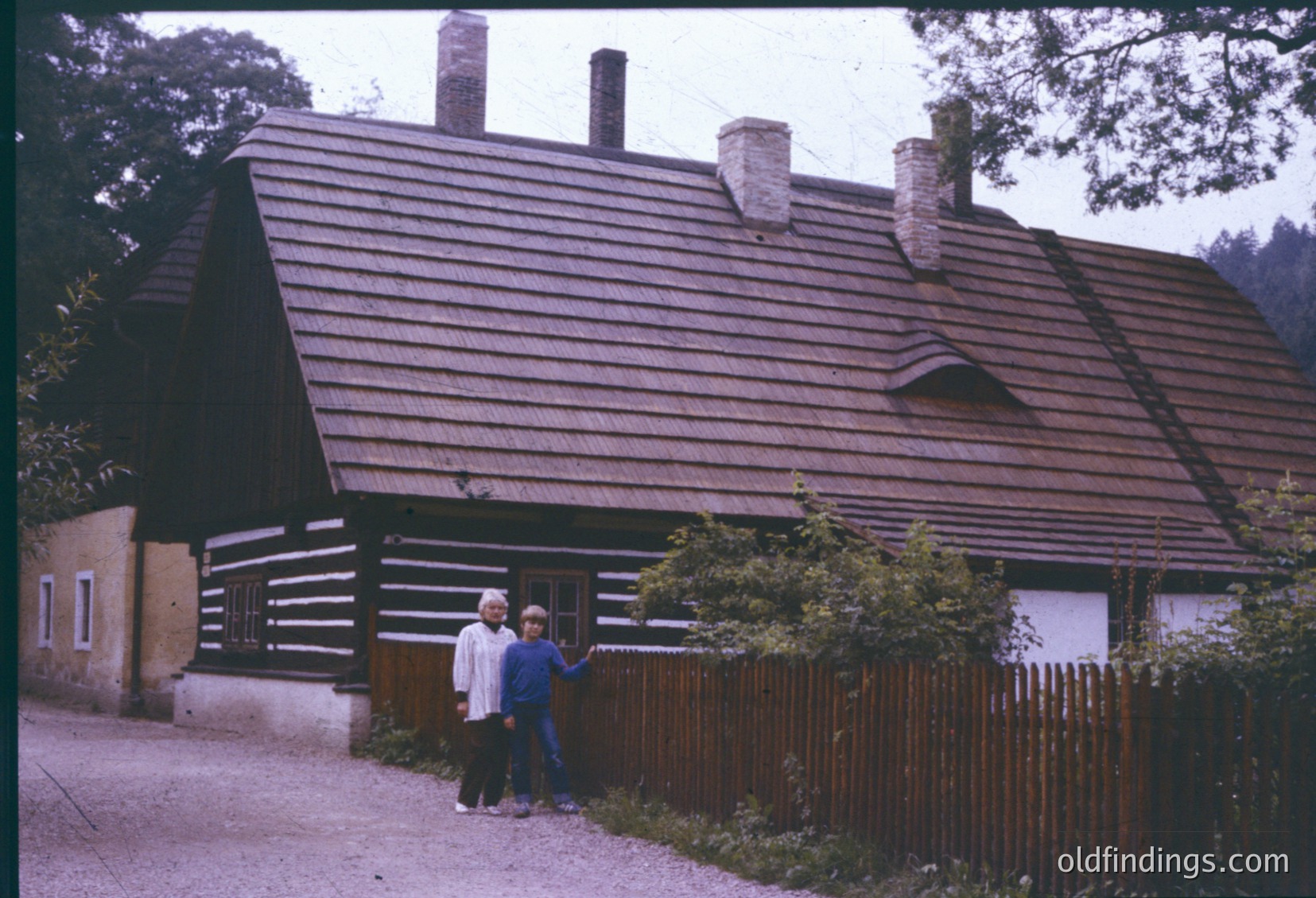Two individuals pose outside a rustic log cabin-style house with a steep, shingled roof and chimneys, framed by a wooden picket fence. The scene suggests a rural European setting, likely from the mid-20th century. [Vintage log cabin exterior with two people posing, rural European architecture, mid-20th century ]