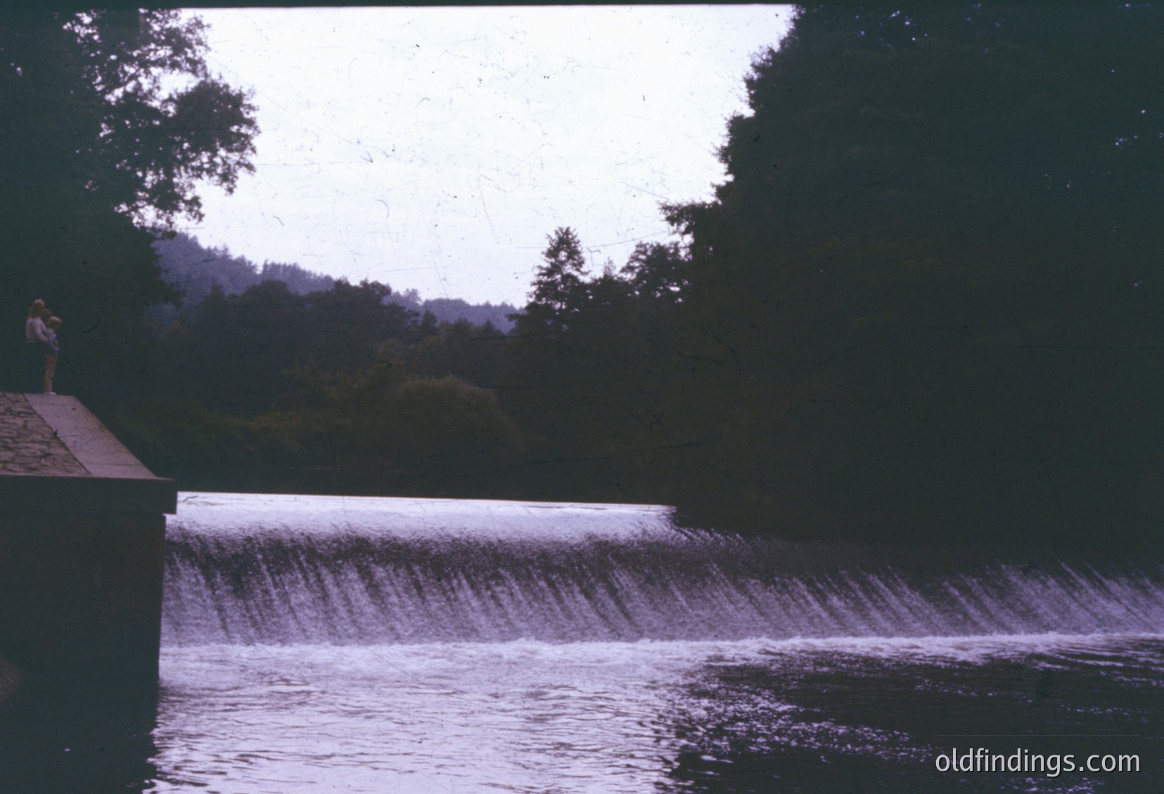 Vintage photo of a concrete dam spillway with cascading water under overcast skies. A lone figure stands on a concrete ledge, silhouetted against the greenery of dense forest. Likely mid-20th century, industrial-era landscape engineering.