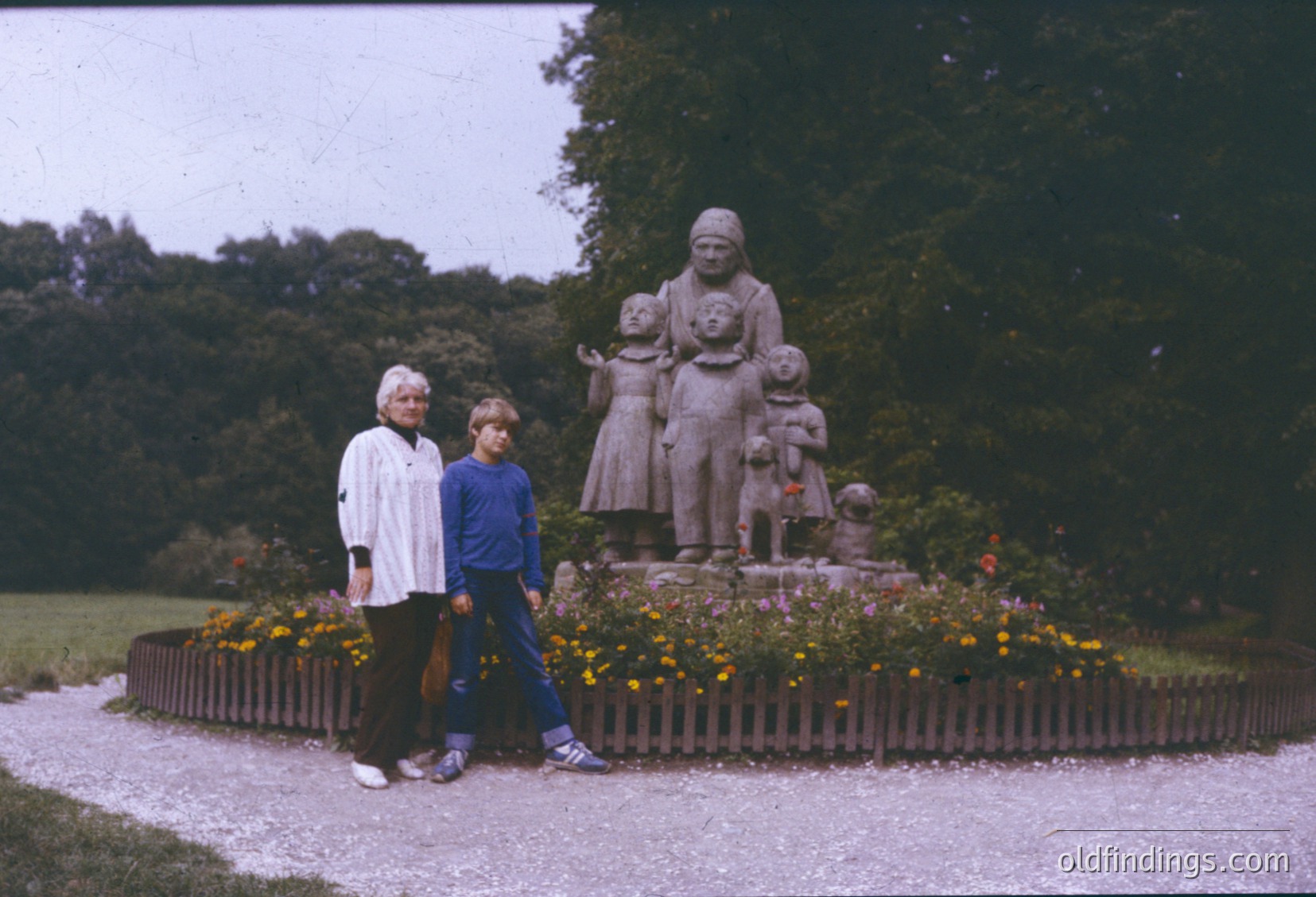 Vintage statue depicting a seated woman cradling three children and a dog, surrounded by vibrant flowers in a landscaped park setting. Two individuals in 1970s-style clothing pose beside the monument. Likely European park, possibly *(Note: Geographic location cannot be confirmed without additional context.)*