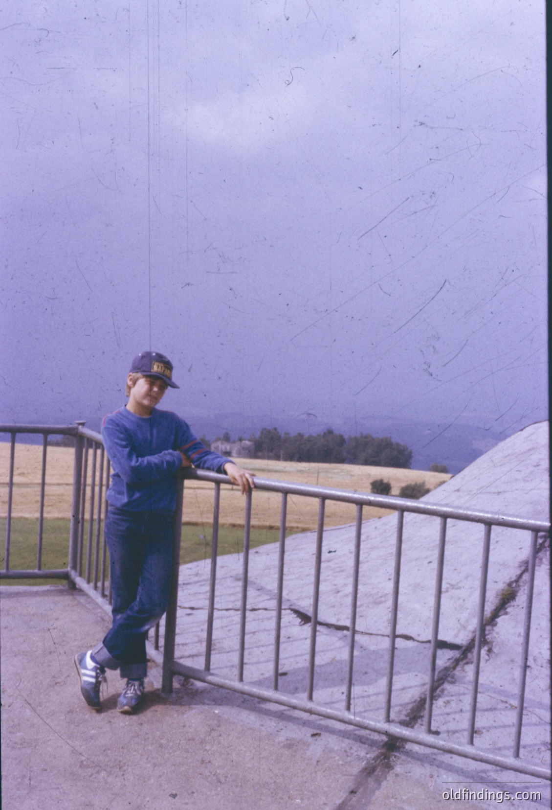 Vintage photo of a young boy in a blue cap and matching tracksuit posing on a concrete balcony overlooking a misty, hilly landscape. The railing and worn concrete suggest mid-20th-century architecture.
