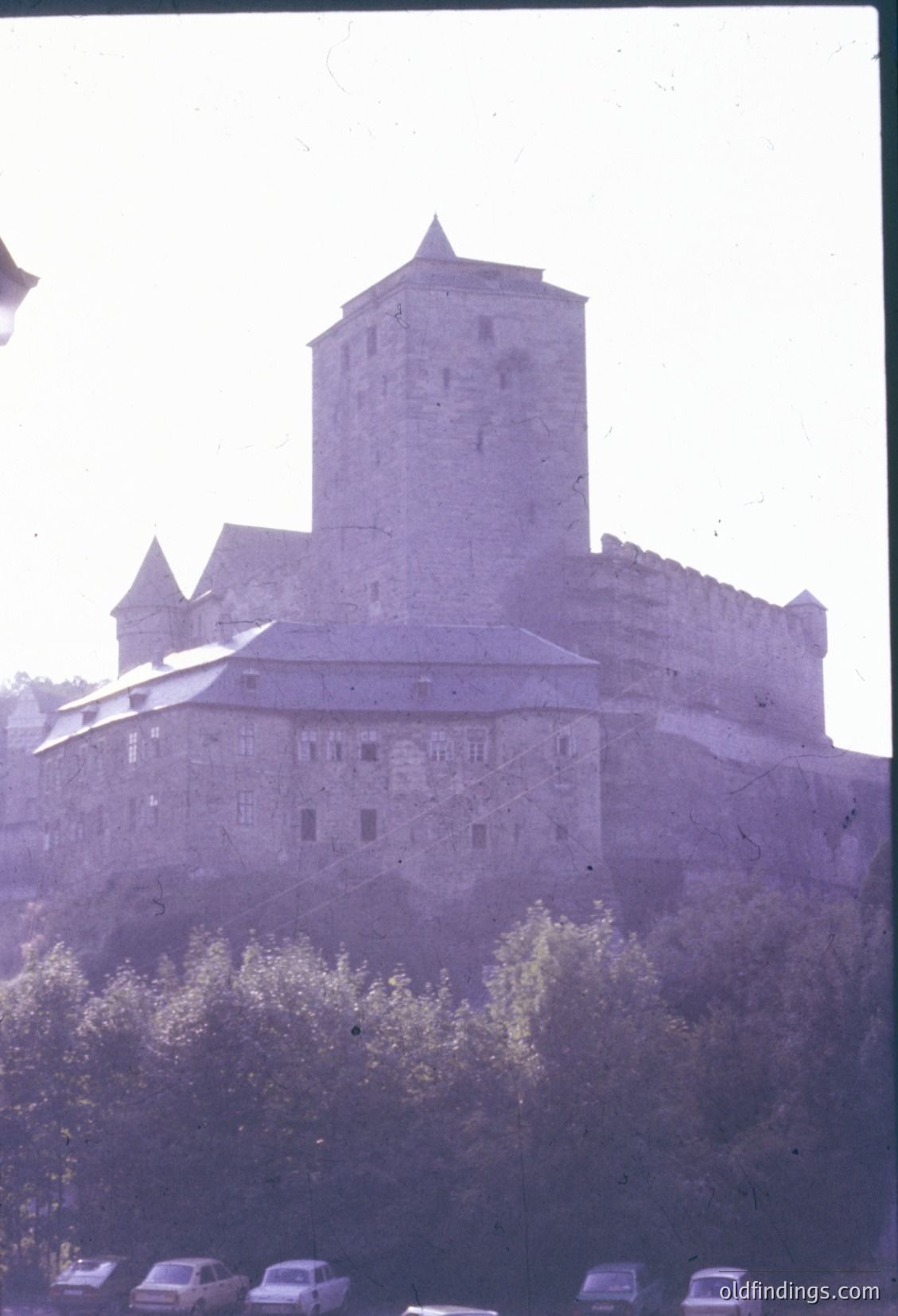 Medieval stone fortress with cylindrical tower and crenellated walls, likely European. Foreground shows vintage cars and trees, suggesting mid-20th century. Architectural style hints at 13th–15th century origins.