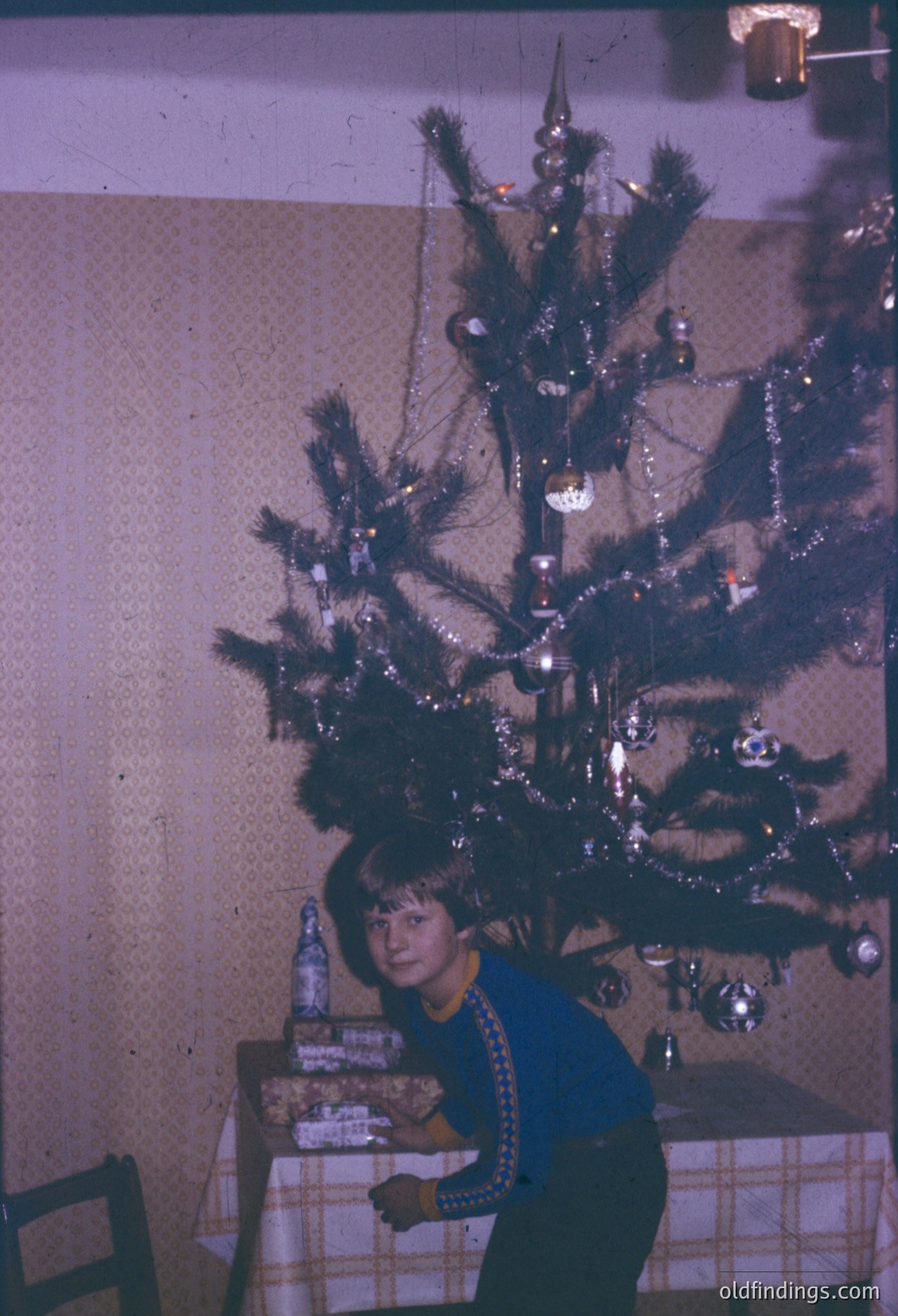 Vintage Christmas scene featuring a young girl seated beside a modestly decorated pine tree adorned with tinsel, baubles, and a single star ornament. She wears a blue sweater with a geometric pattern and holds a wrapped gift. The setting appears to be a modest indoor space, likely a 1960s-1970s home.