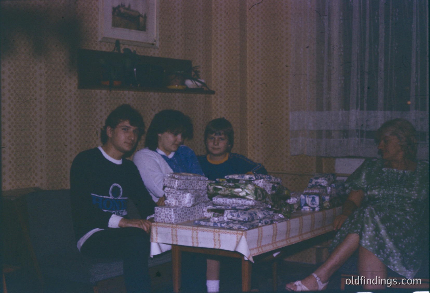 Vintage indoor gathering featuring four adults seated around a table laden with wrapped gifts. Men wear 1970s-style sweatshirts; women in floral dresses. Shelves display ceramic figurines. Warm, dim lighting suggests a cozy, intimate celebration.