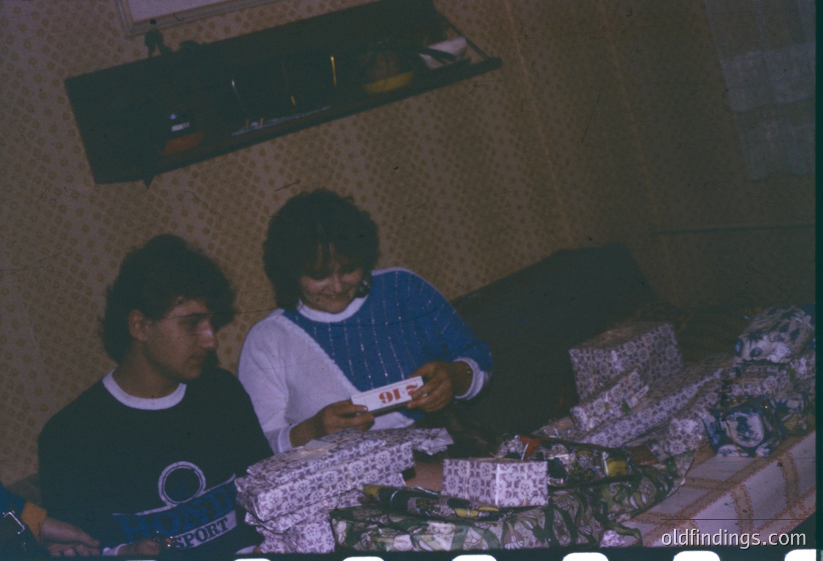 Vintage indoor scene featuring three individuals playing cards on a bed with floral-patterned bedding. The man on left wears a dark turtleneck with a visible "Sport" logo. The woman in center holds playing cards, dressed in a light-colored blouse with a choker necklace. The room’s wallpaper displays a subtle geometric pattern. Likely late 20th century (1970s–1980s), possibly Western Europe or North America.