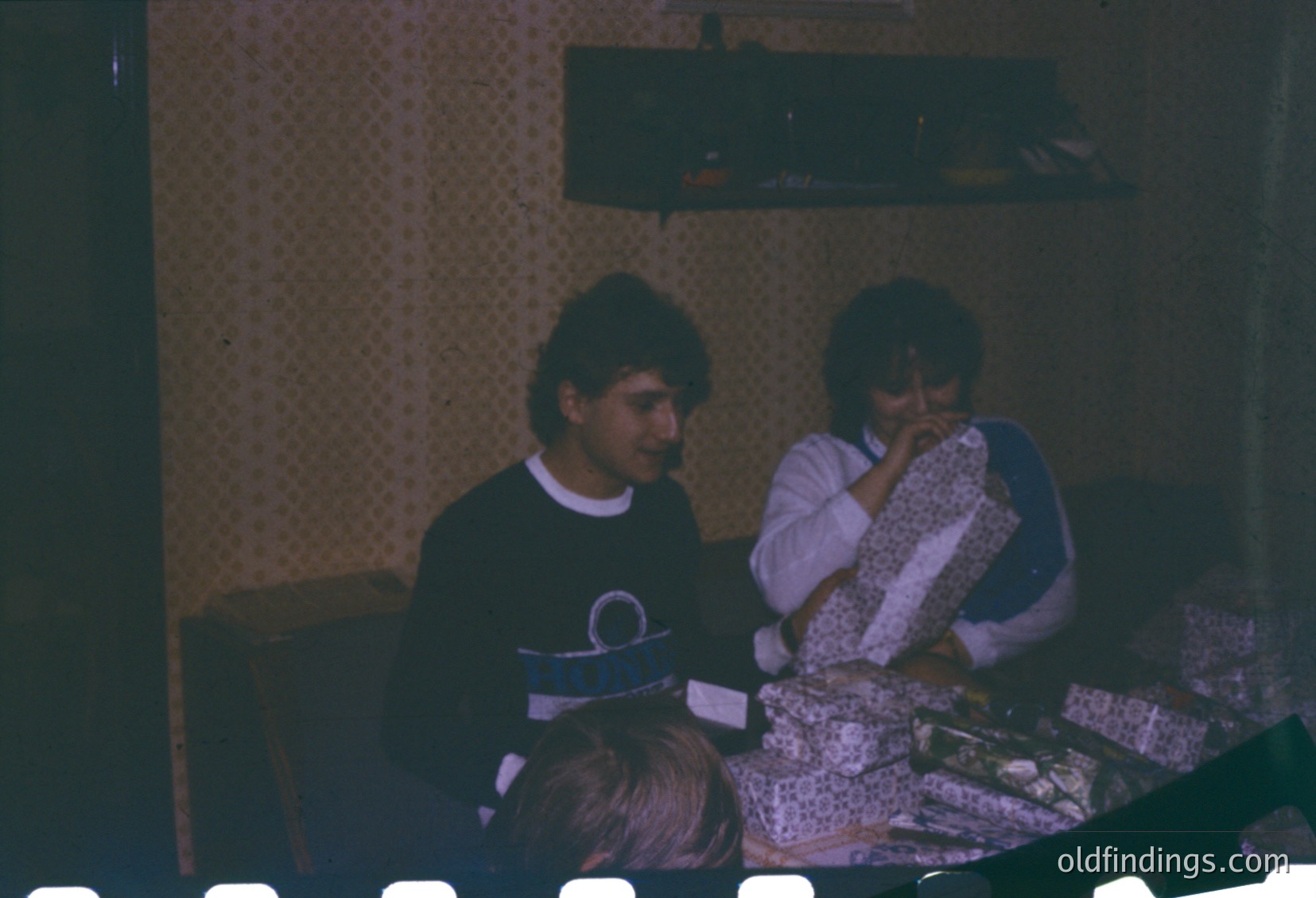 Vintage indoor scene featuring three individuals seated at a patterned table, likely from the 1970s–1980s. The man wears a dark sweater with a logo patch, while the woman holds a patterned cloth. A child’s head is partially visible, suggesting a casual, family gathering. Warm lighting and retro wallpaper enhance nostalgic atmosphere.