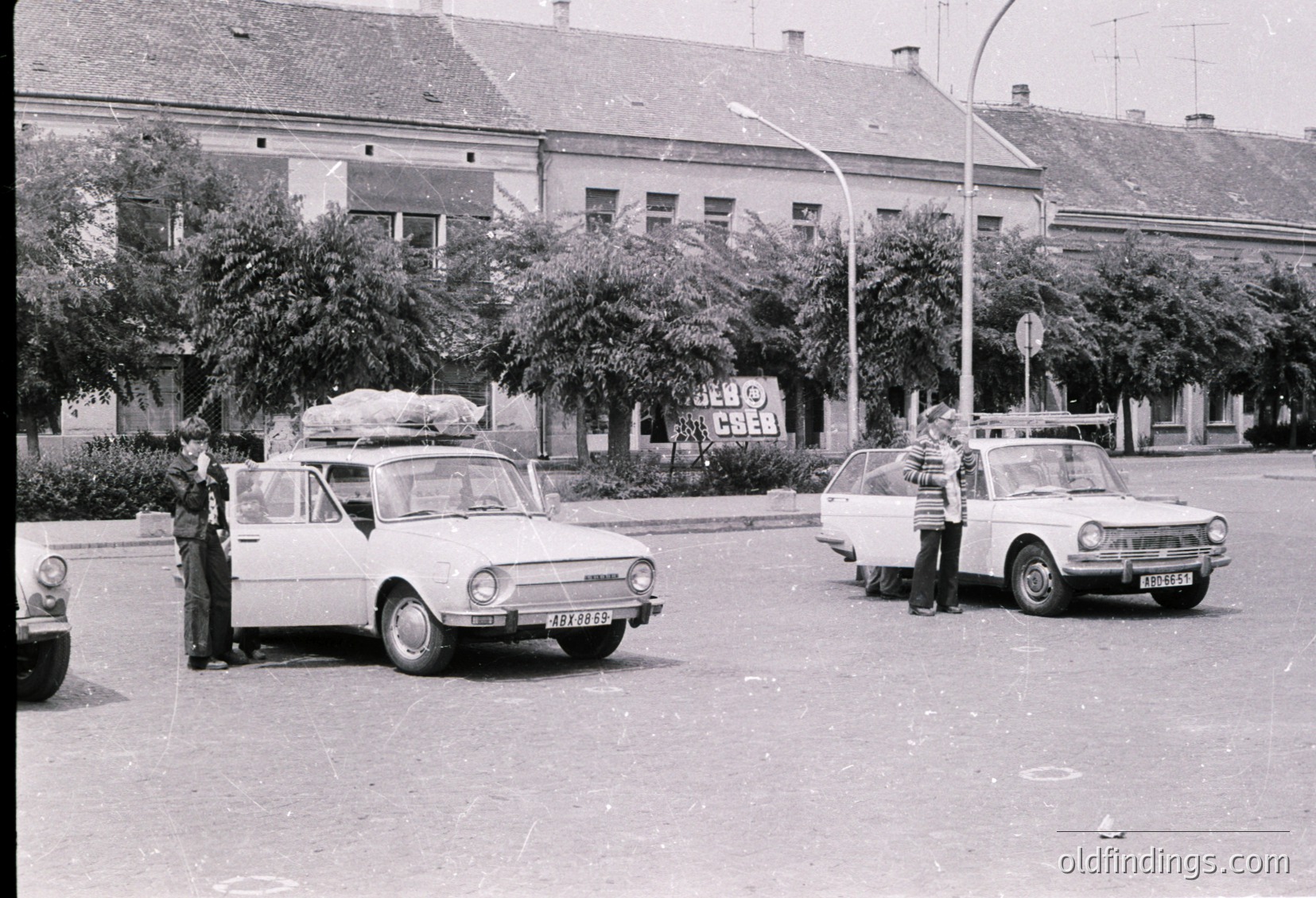 Vintage street scene featuring two Eastern Bloc-era cars (likely Škoda 100/110/120) parked on an urban road. One car has a large white load secured on its roof. Pedestrians in casual 1970s attire stand near vehicles. Soviet-style architecture with multi-story buildings and a "VETO COS" banner in the background suggests a Central/Eastern European setting.