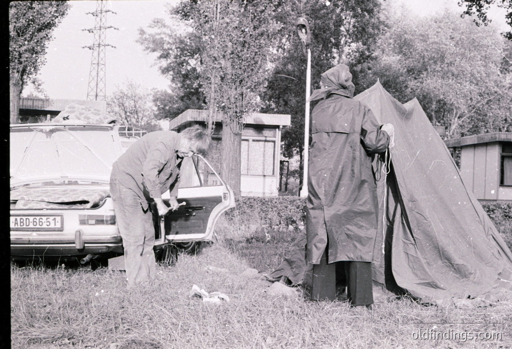 Black-and-white street scene featuring two individuals assisting a woman into a vintage car (ABD 66-51) near a makeshift tent. The man in a light jacket and the woman in a raincoat appear to be in a rural or semi-rural area with trees and a small shed in the background. Likely late 20th century (1970s-1980s).