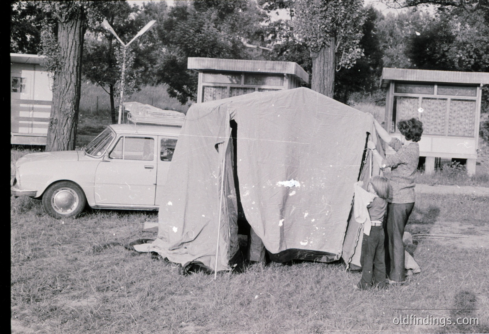 A 1960s-era camping scene: a woman and child setting up a canvas tent beside a vintage car in a grassy area. Nearby, a bus shelter and trees frame the outdoor setting.