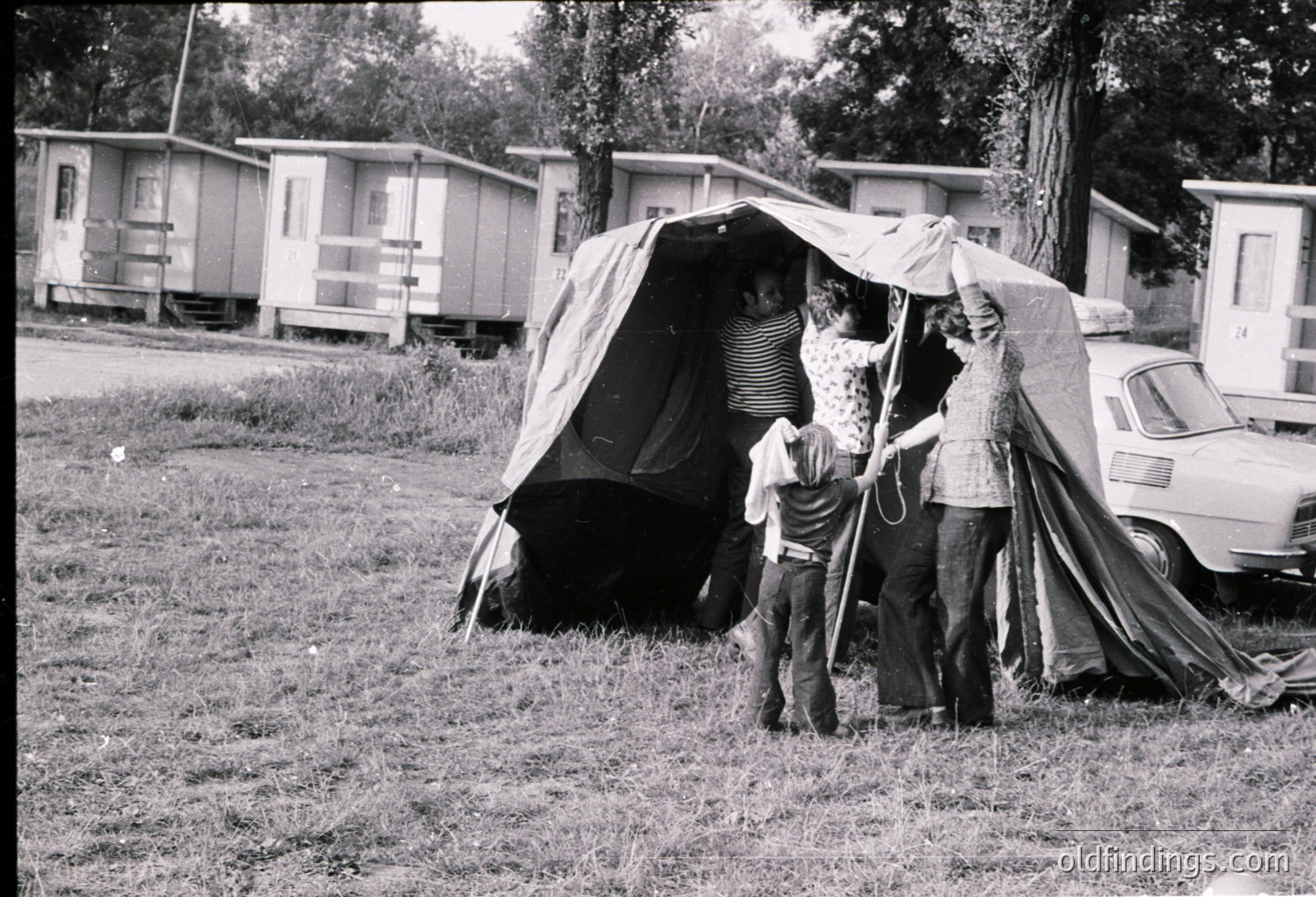 Mid-20th century family camping scene in a grassy area with mobile homes in background. Three adults and a child assist setting up a large canvas tent. Classic vintage car parked nearby.
