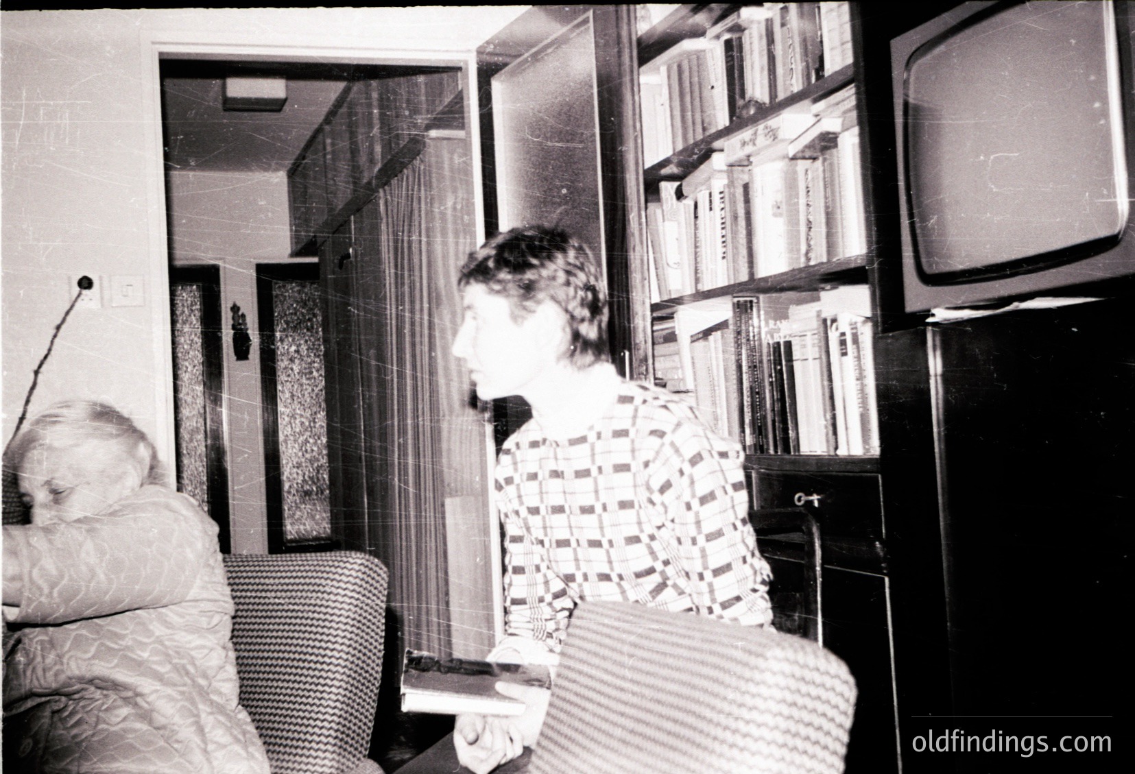 Vintage black-and-white interior shot of a mid-century living room. A woman in a patterned blouse sits on a woven armchair, holding a small dog. Behind her, a bookshelf filled with bound volumes and a bulky CRT TV frame the scene. Classic 1960s–70s home decor with retro furnishings.