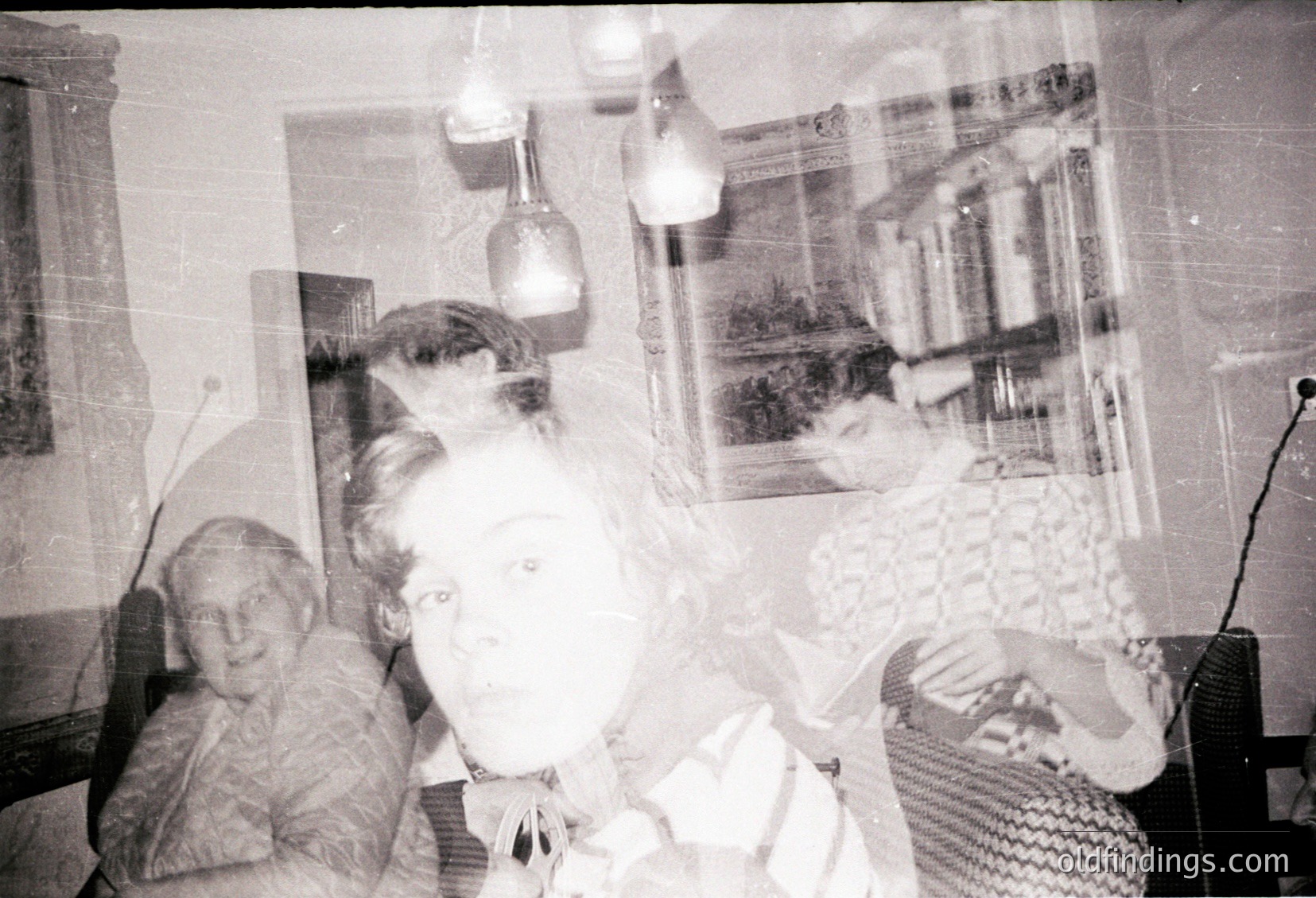 Vintage black-and-white photo of two women in a dimly lit indoor setting, likely mid-20th century. One woman sits on a chair, the other stands beside her, both facing the camera. Framed artwork and a radio on a table suggest a lived-in, domestic space. Textured wallpaper and a lamp with a white shade add to the nostalgic atmosphere.
