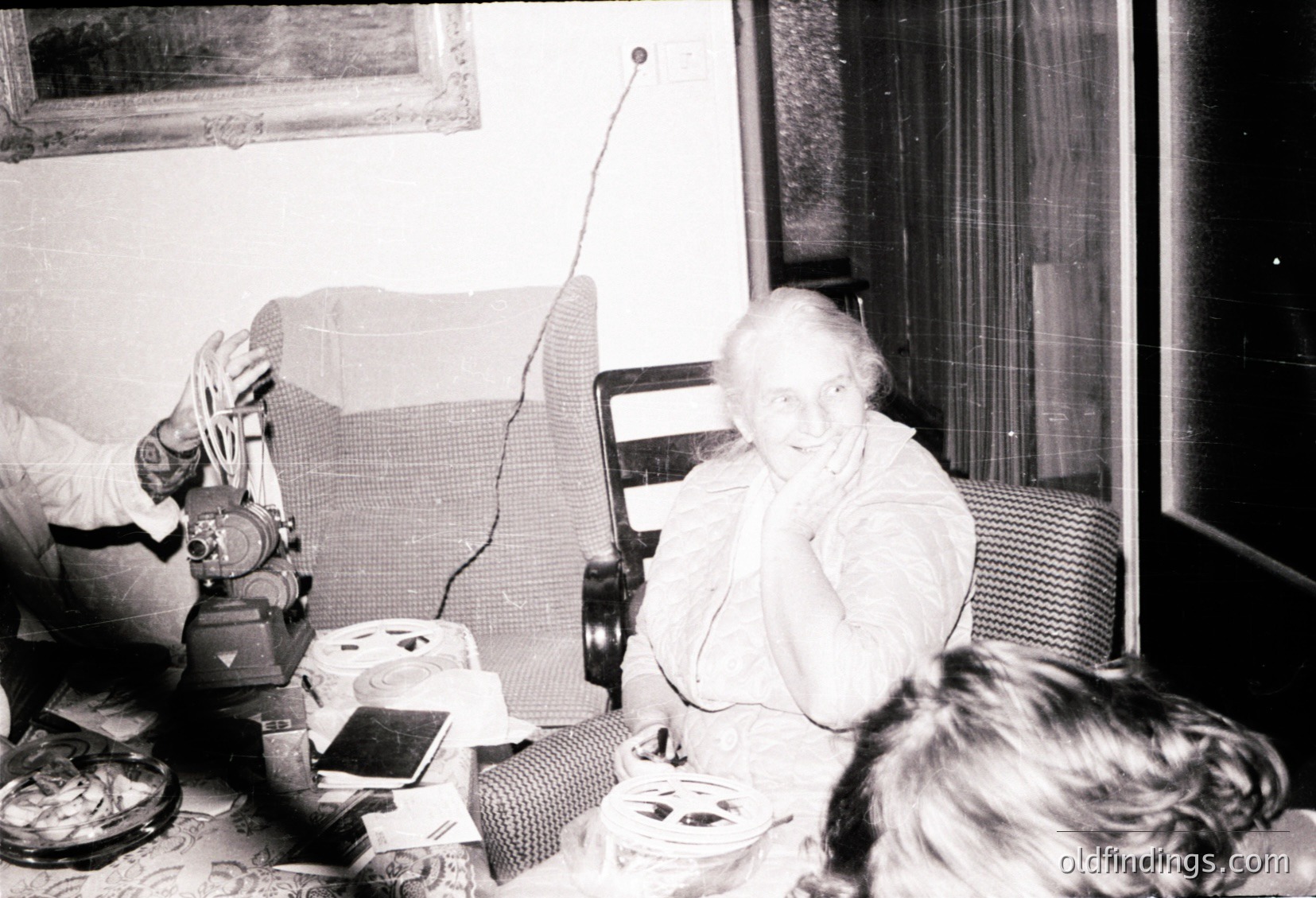 Vintage black-and-white interior shot of an elderly woman seated in a patterned armchair, holding a small object (likely a phone or device). Surrounding her: a vintage radio on a side table, a cluttered coffee table with dishes, and a child’s arm in foreground. Mid-20th century domestic setting, likely 1950s–1960s.