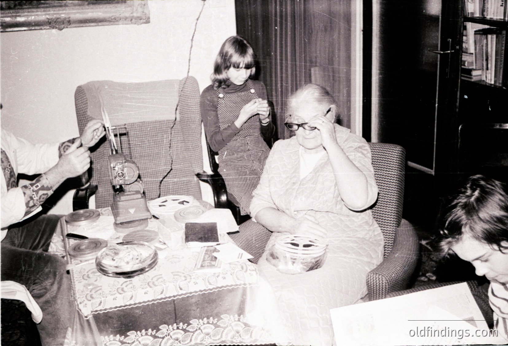 Vintage black-and-white indoor scene featuring a seated elderly woman in glasses holding her face, surrounded by three children. Tabletop displays mid-20th-century decor: vintage radio, patterned tablecloth, and a cake. Bookshelves and armchairs suggest a lived-in, familial home setting.