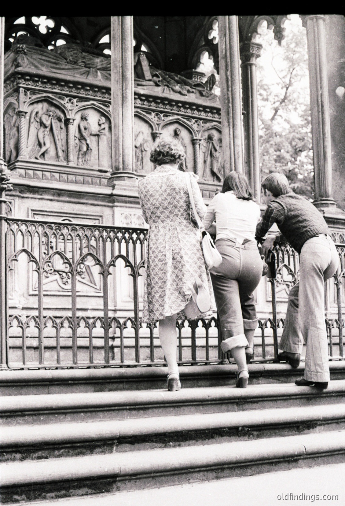 Black-and-white photo of three individuals ascending ornate stone steps in a historic European setting, likely 1960s–1970s. Intricate Gothic-style railings and relief carvings depict human figures. The woman in a floral dress and men in wide-legged trousers suggest mid-century fashion.