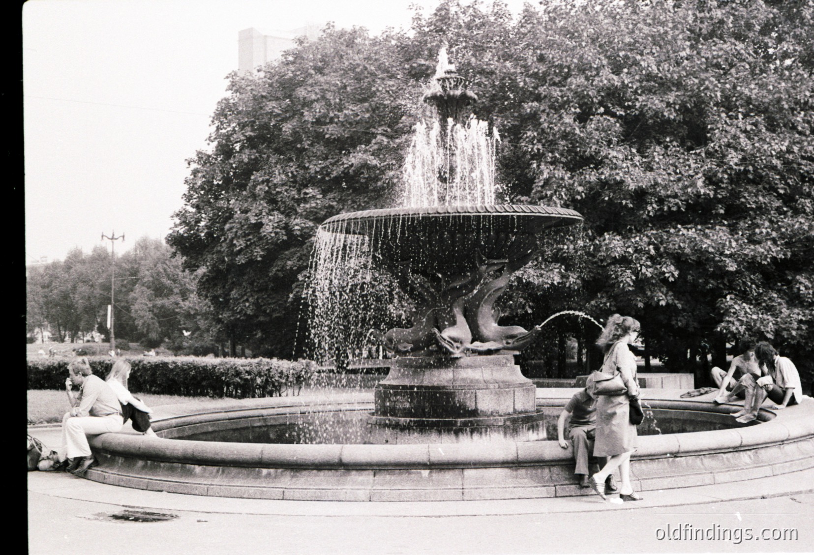 Classic mid-20th-century fountain with sculpted sea creatures, surrounded by lush greenery. People in vintage attire—men in suits, women in dresses—relax around the base. Urban park setting, likely European.