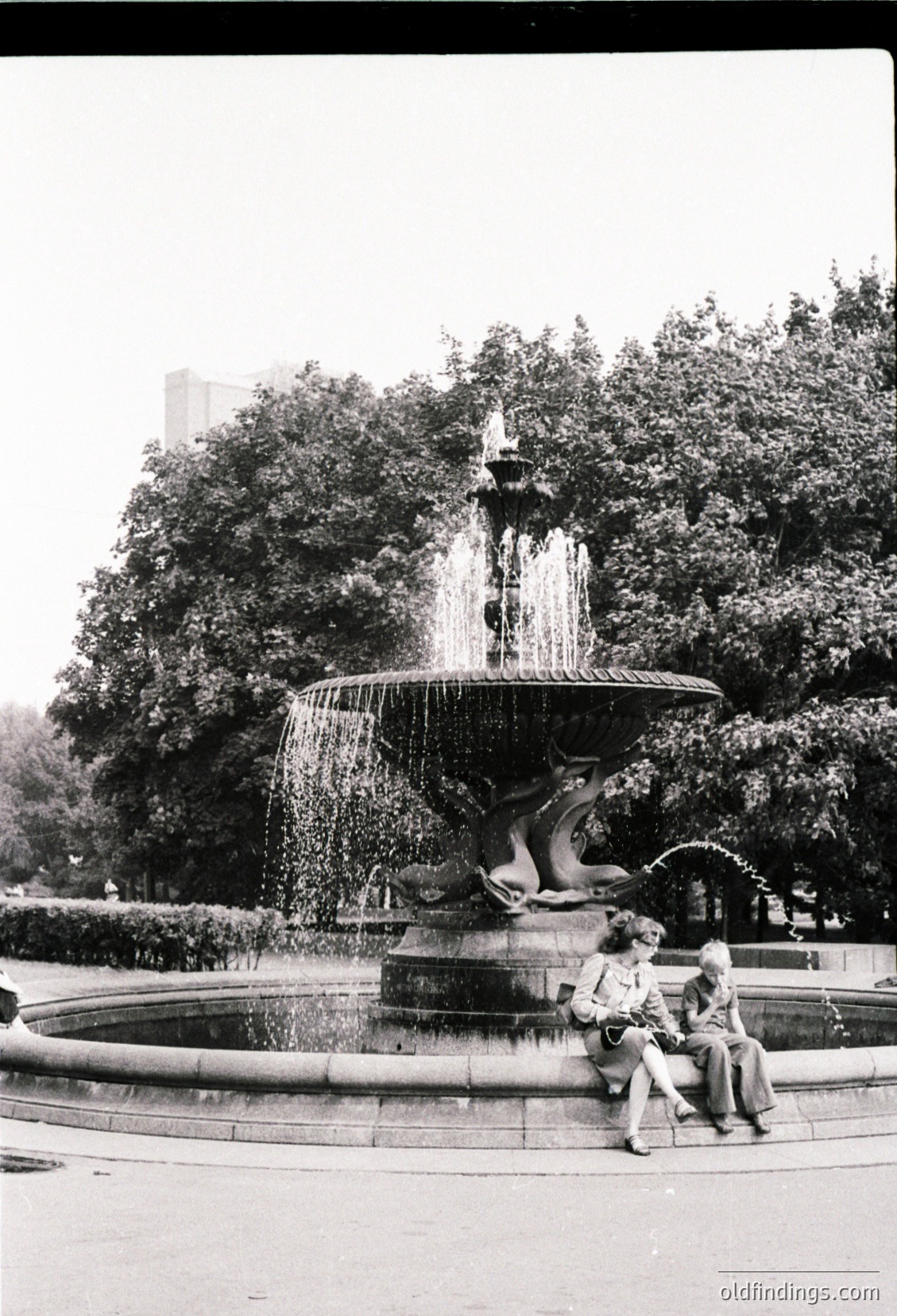 Vintage black-and-white shot of a multi-tiered fountain with sculpted sea creatures, likely from the 1950s–1960s. Two seated figures rest on the fountain’s edge, surrounded by lush greenery and urban trees. Architectural details suggest a European city park.