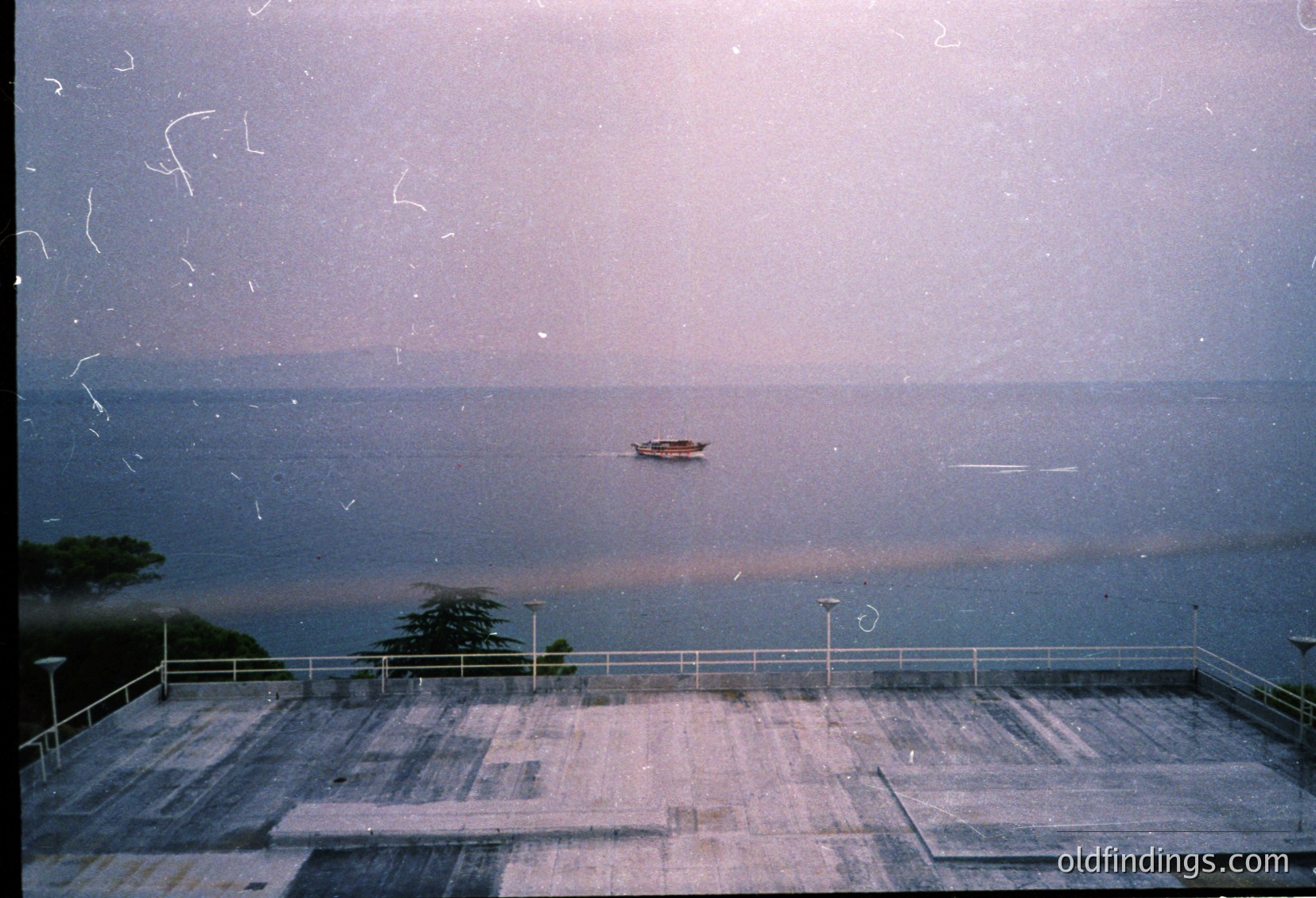 Vintage seaside view through a slightly scratched window, showing a lone boat on calm waters. Concrete balcony with metal railings and scattered debris. Coastal landscape with distant trees and horizon. Likely Mediterranean or Black Sea region, 1980s–1990s.