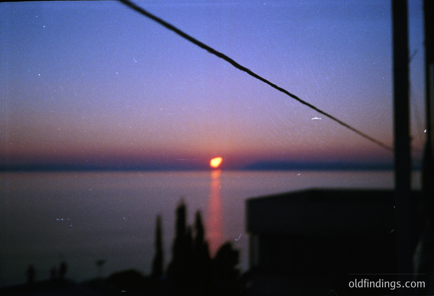 Vintage-style shot of a sunrise/sunset beam through a window frame, silhouetted by silhouetted trees and a faint coastal horizon. Warm light filters through a slightly blurred lens, creating a nostalgic, cinematic effect. Likely late 20th century, possibly or .