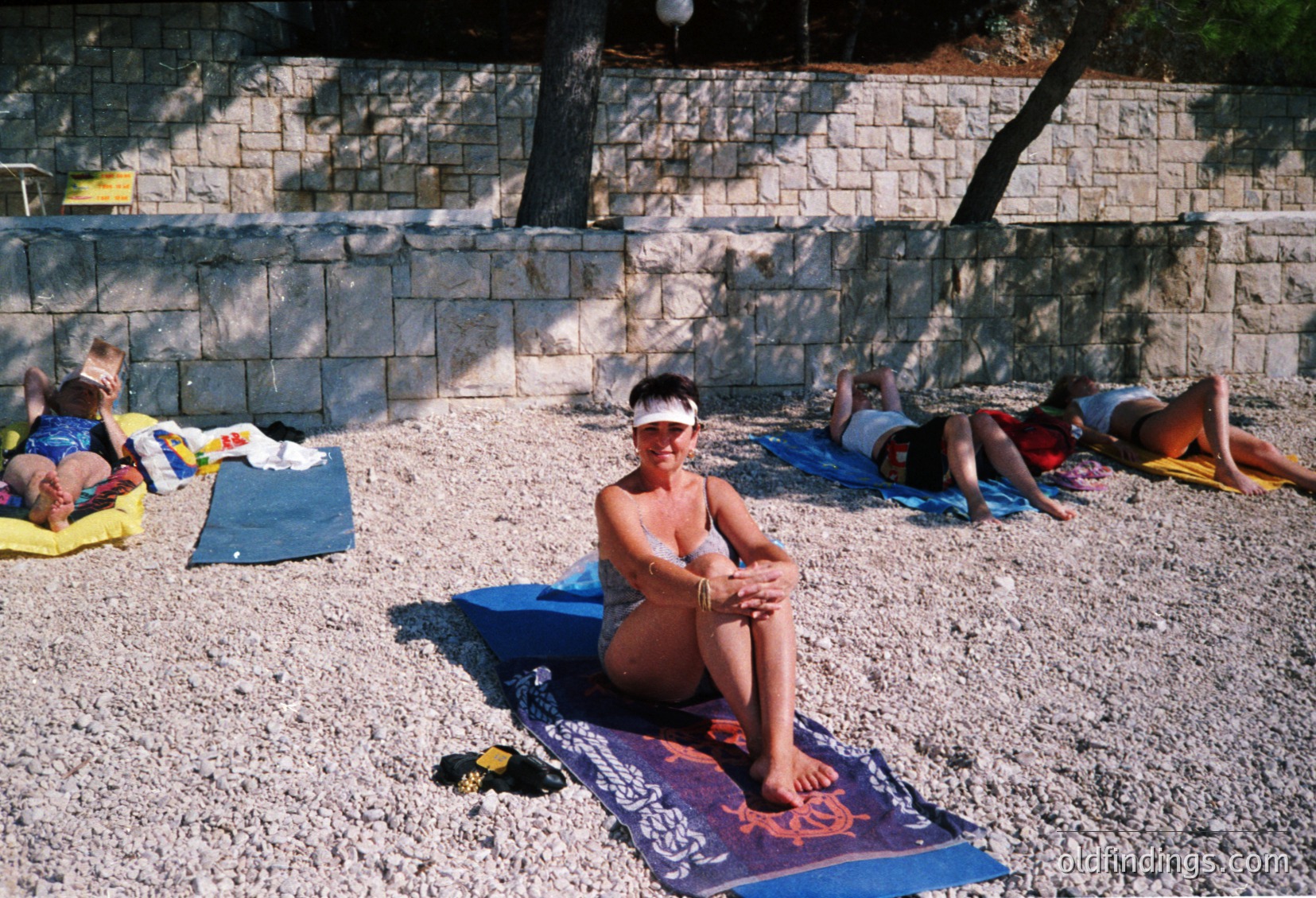 Sunbathing scene on a pebble beach with stone retaining wall. Six people lounging on towels/mats, one woman in foreground wearing sunglasses and bracelets. Mid-20th century beachwear and relaxed posture suggest leisure culture. Likely Mediterranean coastal area.