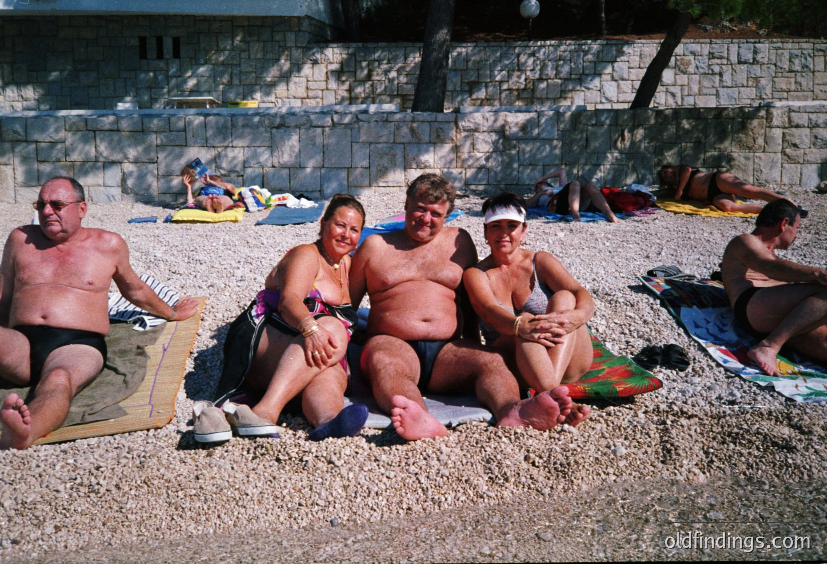 Group of five adults lounging on a pebble beach, mid-20th century. Four seated on towels, one standing beside them. Stone retaining wall and concrete structures in background suggest a resort setting. Casual summer attire reflects 1970s-1980s beach culture.