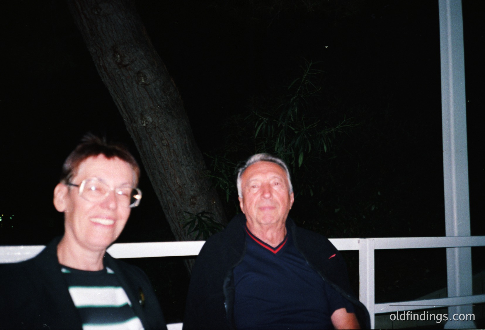 Two adults pose outdoors at night under artificial lighting, framed by a tree trunk and metal railing. The man wears a dark jacket with red trim, while the woman wears glasses and a striped shirt. Style suggests mid-2000s casual wear.