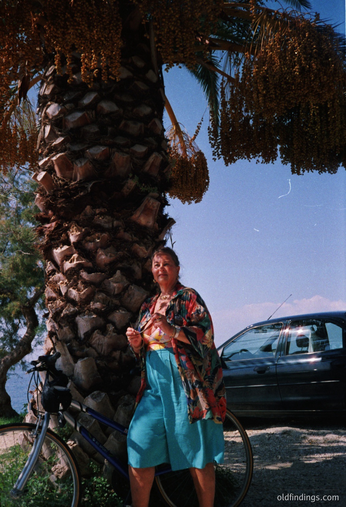 A person in a patterned shirt and turquoise pants sits atop a stacked stone wall, mirroring their reflection in a calm body of water. A bicycle leans against the wall beside them, while a dark vehicle and tree foliage frame the scene. Likely a 1990s–2000s outdoor setting, possibly a park or lakeside area.