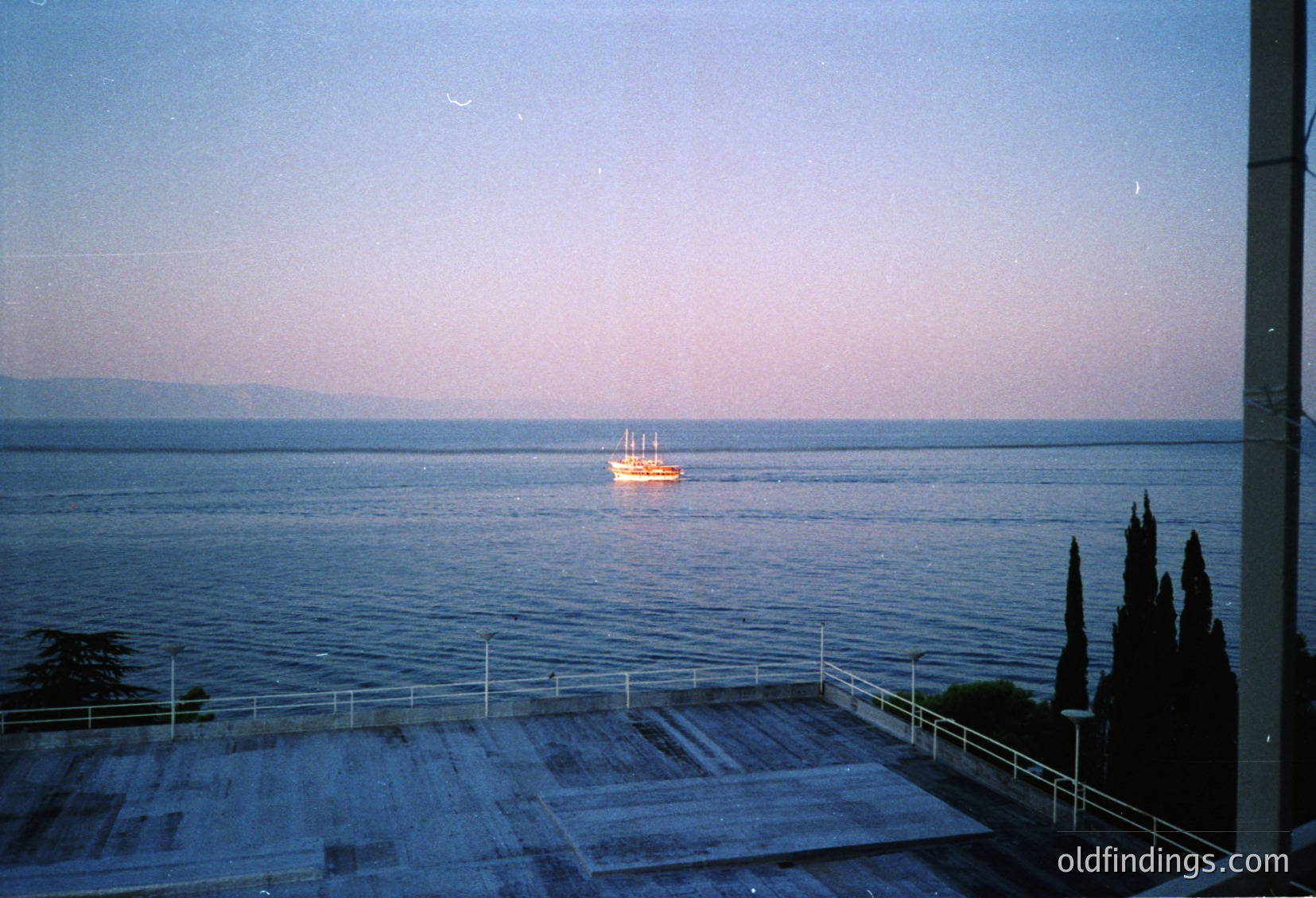 Vintage seaside balcony view with a lone sailboat on calm waters at dusk. Concrete railing and potted cypress trees frame the scene, suggesting a coastal resort or hotel. Soft pastel sky transitions to deepening blue over the horizon. Likely Mediterranean or Black Sea region, 1970s-1990s.