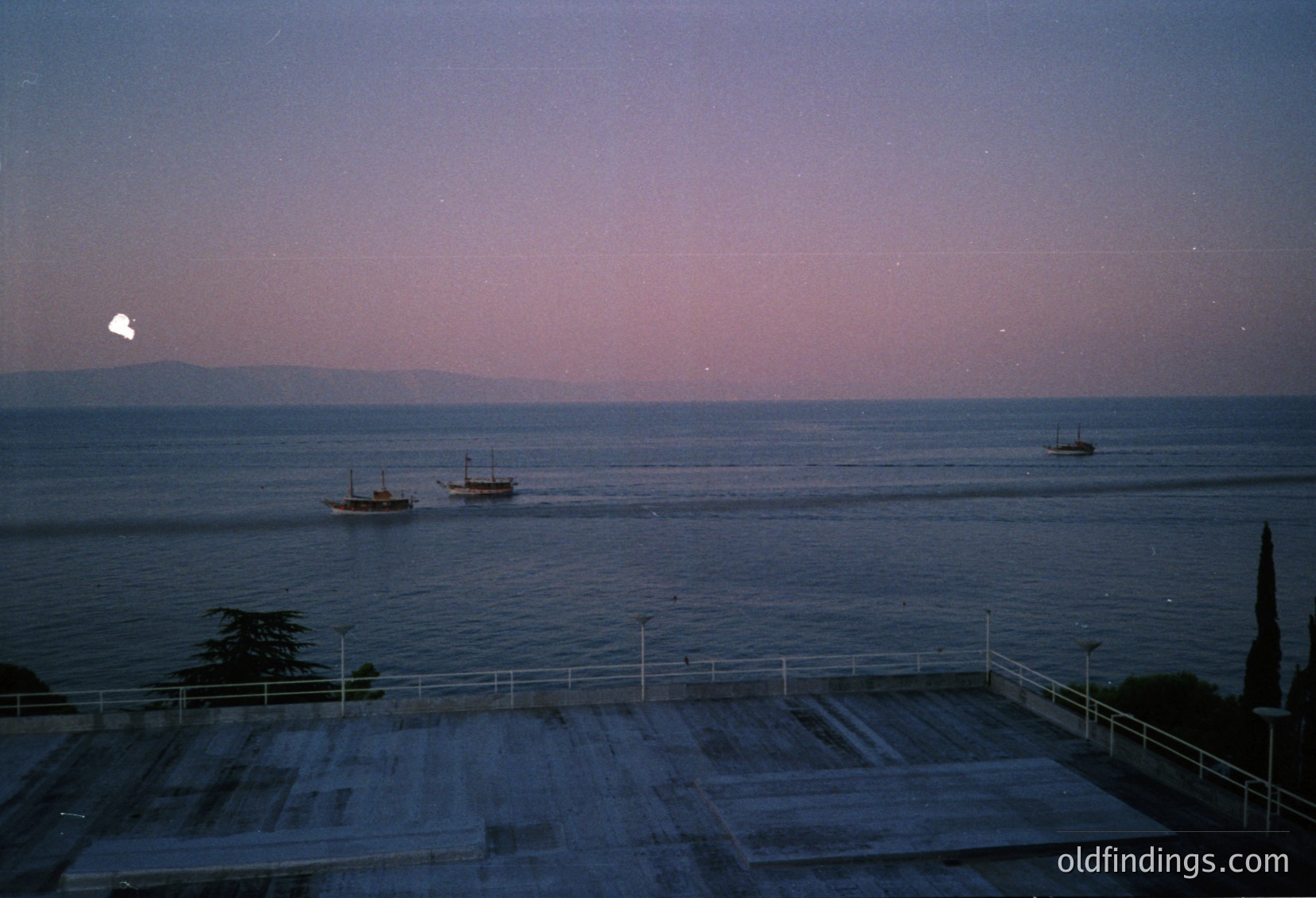 Twilight seascape with three small wooden boats anchored near shore. Concrete terrace with metal railings in foreground. Distant silhouette of landmass under fading light. Likely coastal Mediterranean or Black Sea region.