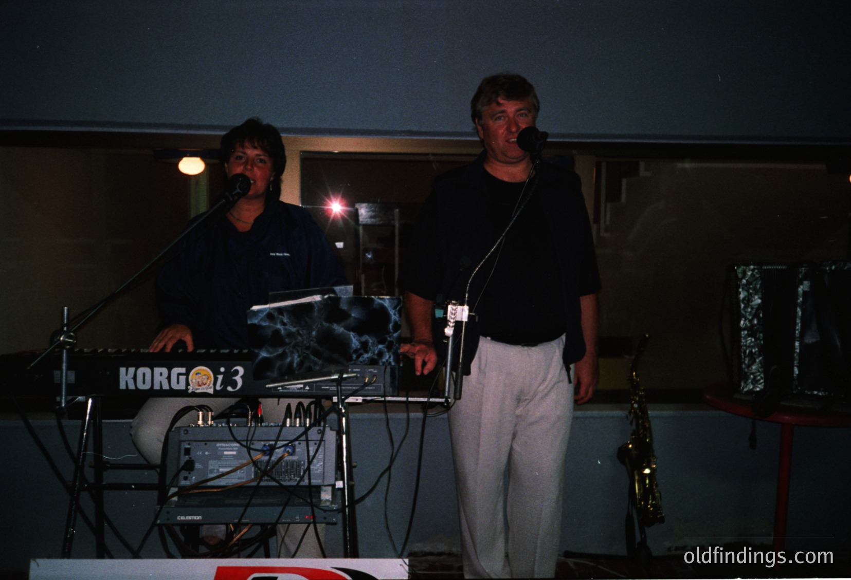 Two musicians performing live on stage, 1980s–1990s era. Left: keyboardist playing a Korg Poly-800i synth, right: saxophonist holding a tenor sax. Stage lighting highlights equipment and performers against a dark backdrop. Indoor concert venue setting.