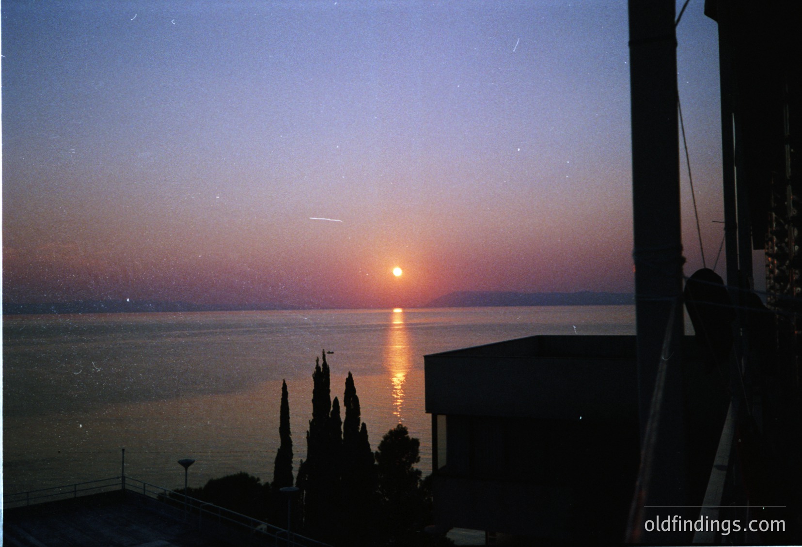 Sunset over a coastal horizon with golden orb reflecting on calm waters. Silhouetted cypress trees frame the right edge, while a building’s rooftop occupies the left. Vintage film grain and slight overexposure suggest mid-20th century photography.