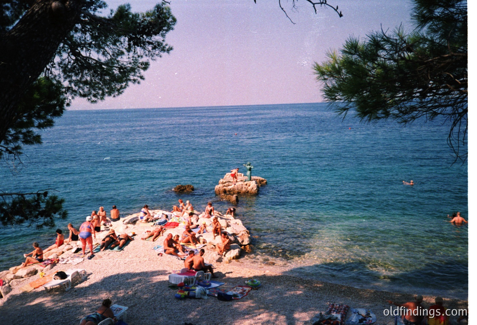 Vintage seaside scene with sunbathers on a sandy beach, framed by pine trees. Bright orange/red swimsuits dominate the crowd, suggesting mid-20th century beach culture. Clear water and shallow entry point visible. Likely Mediterranean or coastal European location.