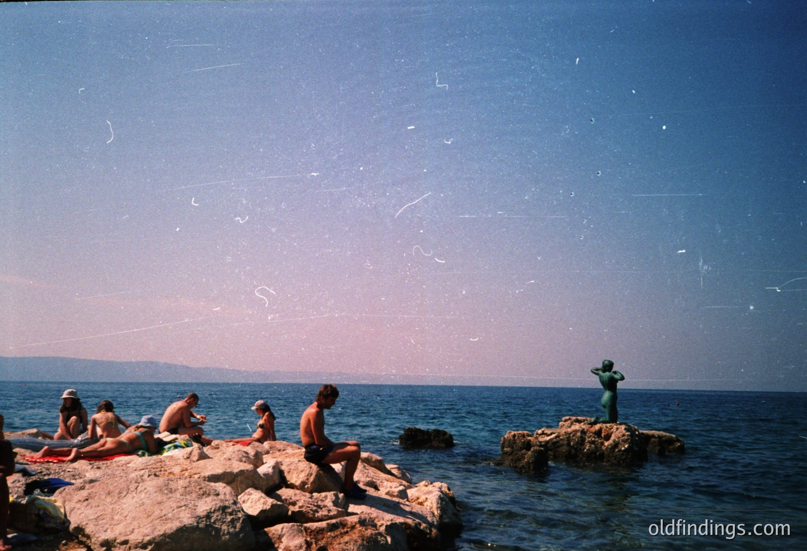 Reflection of people in shallow water, likely a beach or lakeside. Distinctive vintage filter and grain suggest mid-20th century photography. Footwear includes sandals and flip-flops, indicating warm climate.