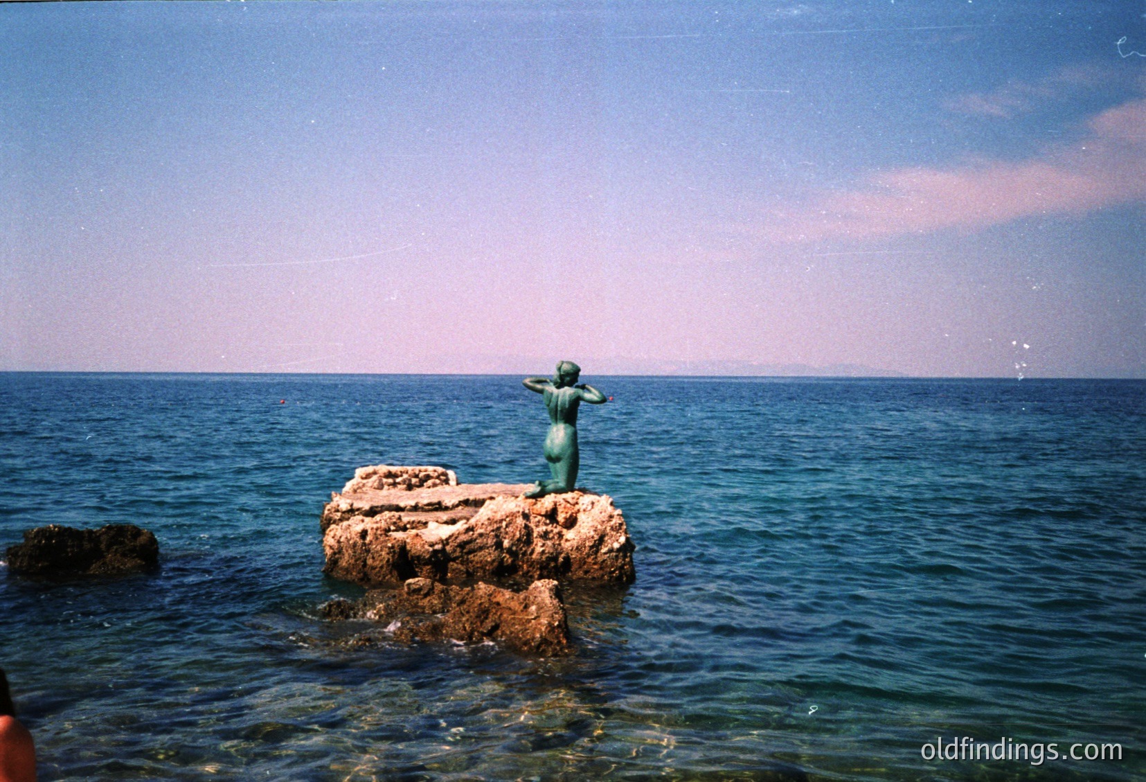 Underwater shot of a submerged stone fountain with a single greenish-blue jet of water. The structure appears weathered, suggesting age or natural erosion. Likely a public pool or historic water feature.