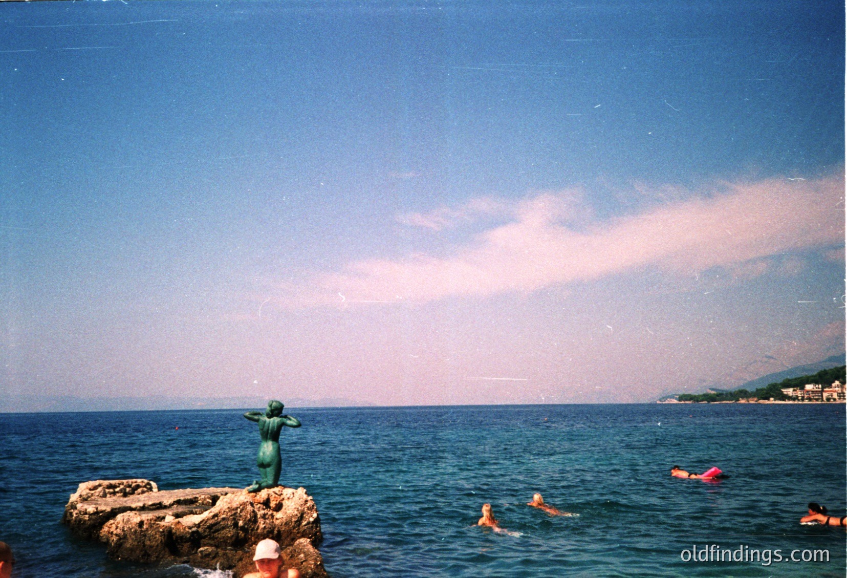Aerial view of a shallow, sandy beach with clear turquoise waters. Three swimmers in the background near rocky formations. Vintage filter suggests mid-20th century coastal tourism vibe.