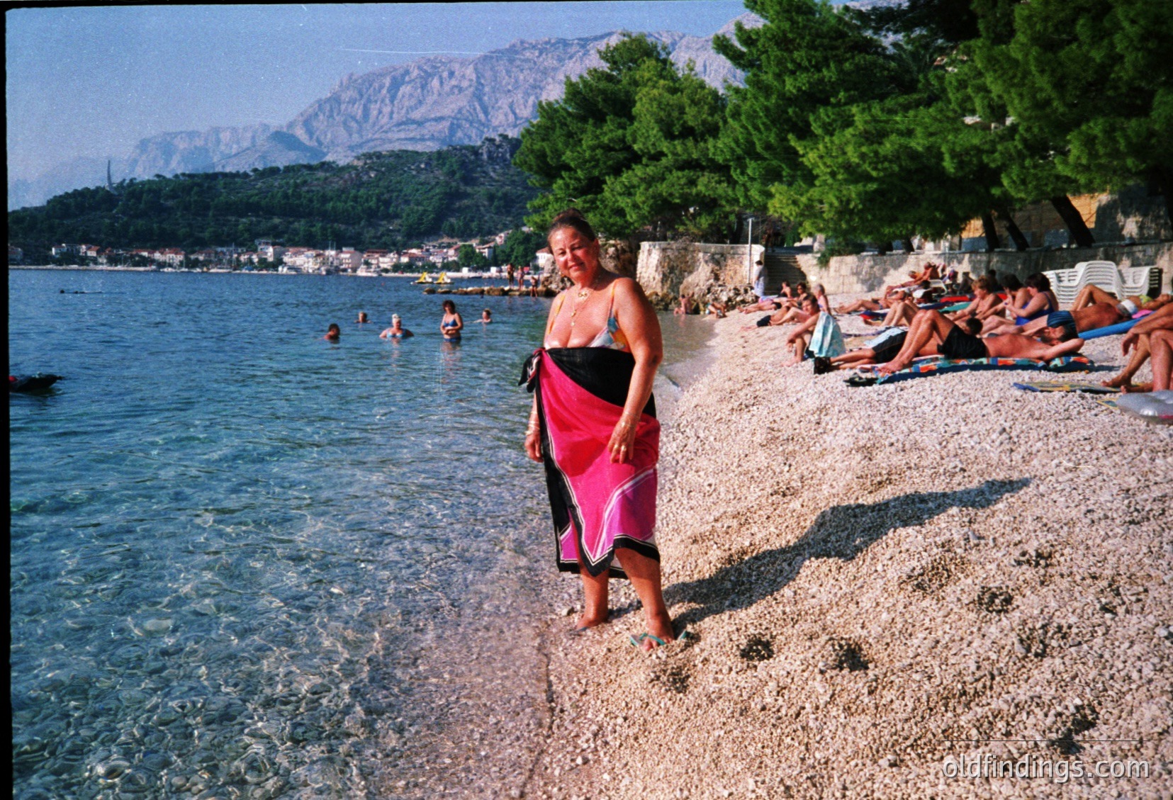A swimmer mid-dive off a concrete pier into clear blue water, surrounded by pebbled shore. Sunlit reflections create a mirror-like effect on the water. Beachgoers relax on towels and chairs in the background, suggesting a coastal resort setting. Likely Mediterranean or Adriatic region, summer season.