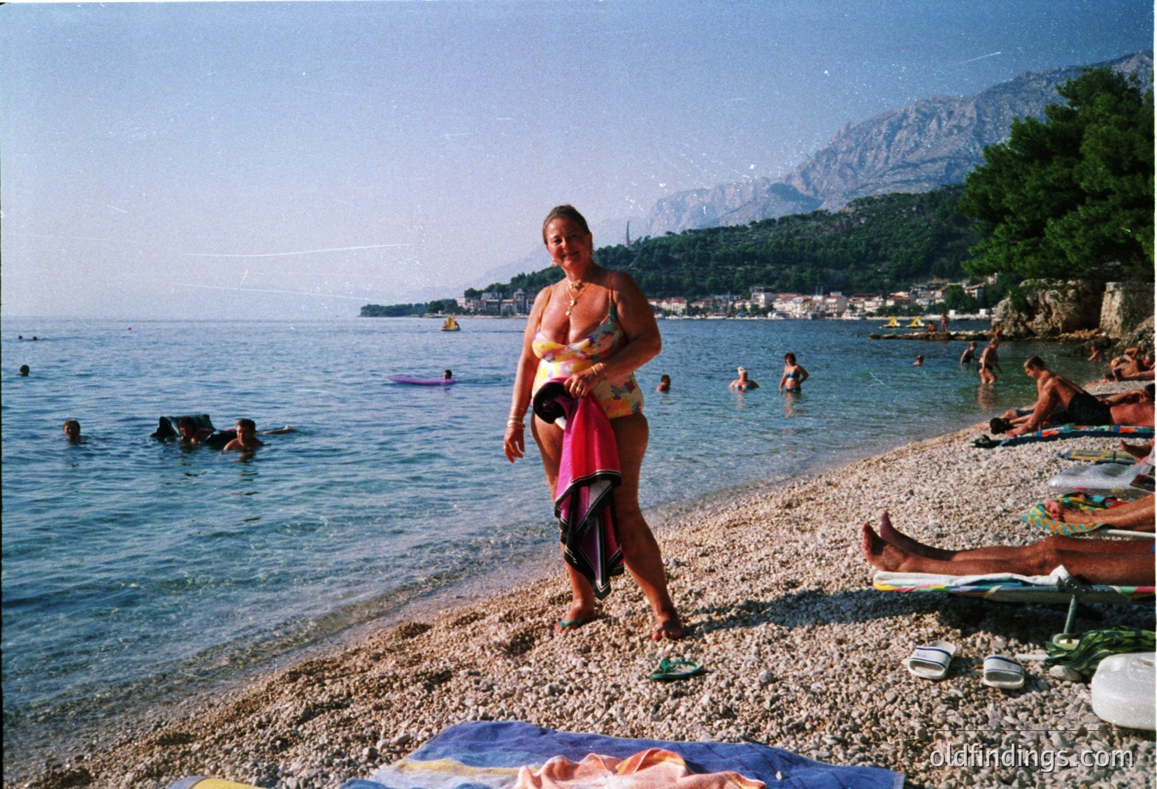 Mid-air dive off rocky cliff into turquoise waters; 1970s beach scene with sunbathers on towels below. Bold red-and-white swimwear contrasts with natural tones. Cliffside lounge chairs and scattered umbrellas suggest a popular seaside spot.