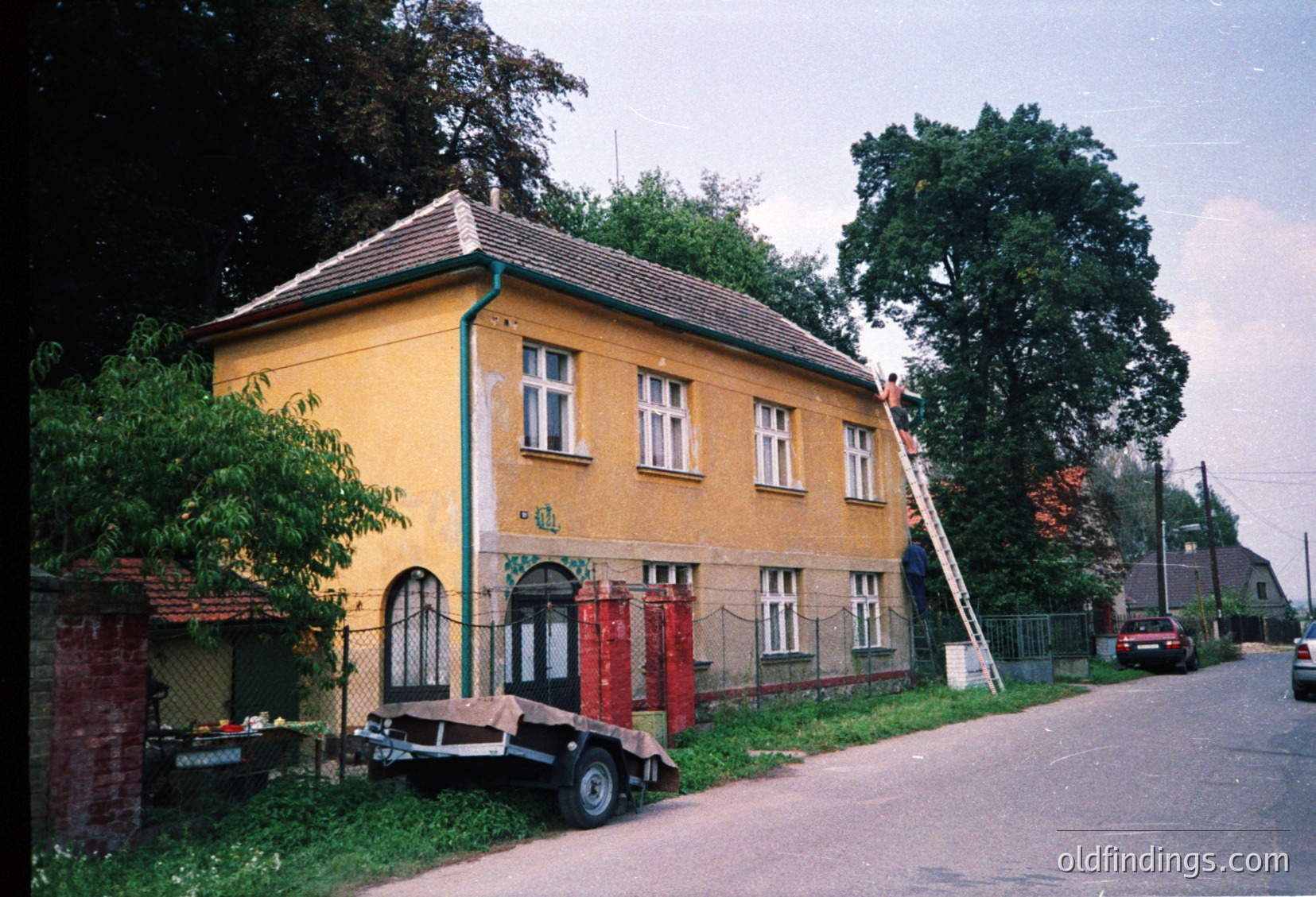 Two-story yellow building with red-tiled roof, likely residential or small commercial, featuring arched entrance and white-framed windows. Ladder leans against upper wall, suggesting maintenance or renovation. Red fire escape ladder mounted on exterior. Trailer parked in front with miscellaneous items. Overgrown greenery and chain-link fence. Street scene with parked vehicles and trees.
