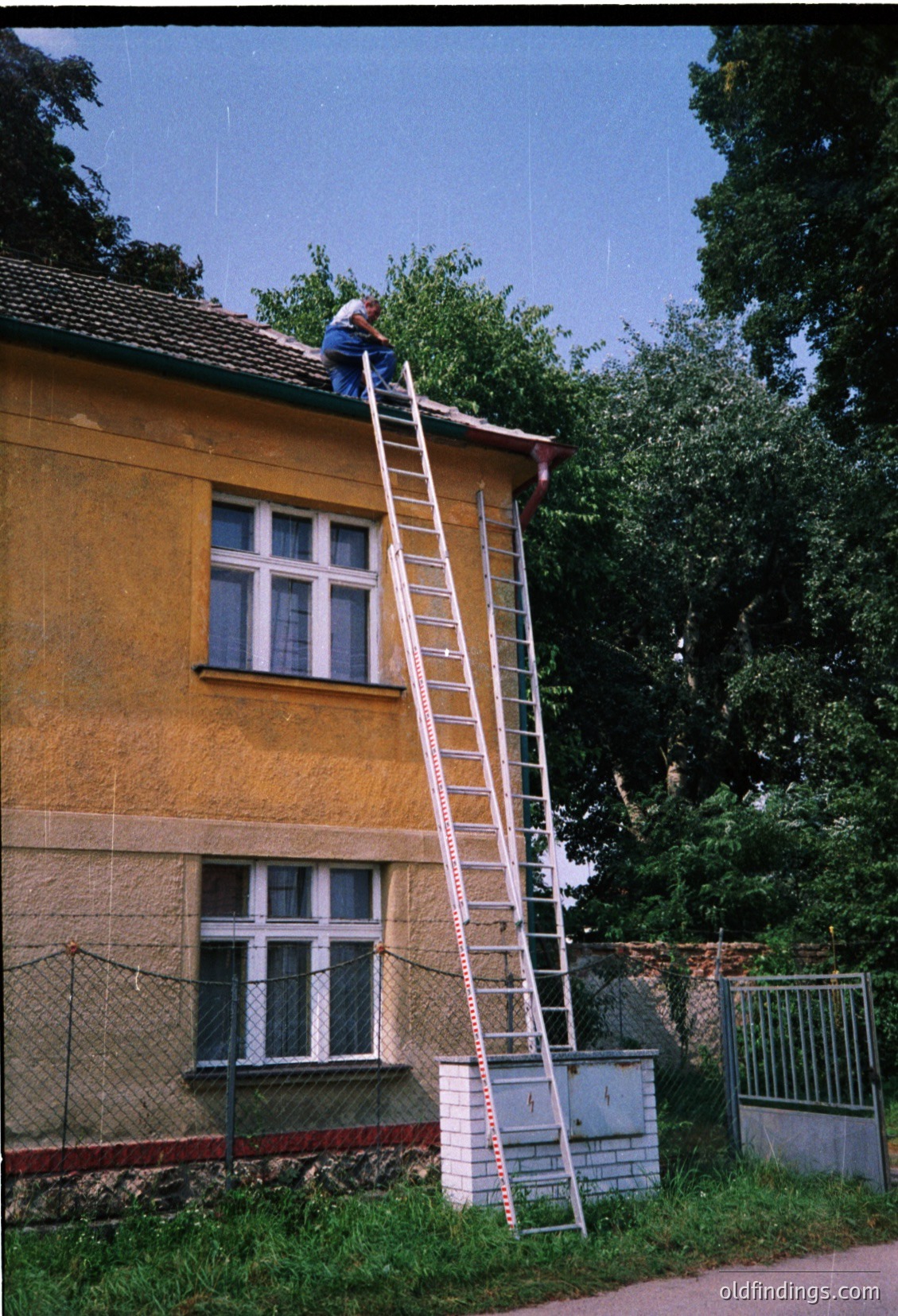Aerial view of a mid-rise building under maintenance, featuring a white extension ladder against a yellowish stucco facade. Two workers in safety gear (helmets, harnesses) appear on the rooftop. Surrounding foliage and a chain-link fence frame the scene. Likely 1990s–2000s urban/suburban setting.