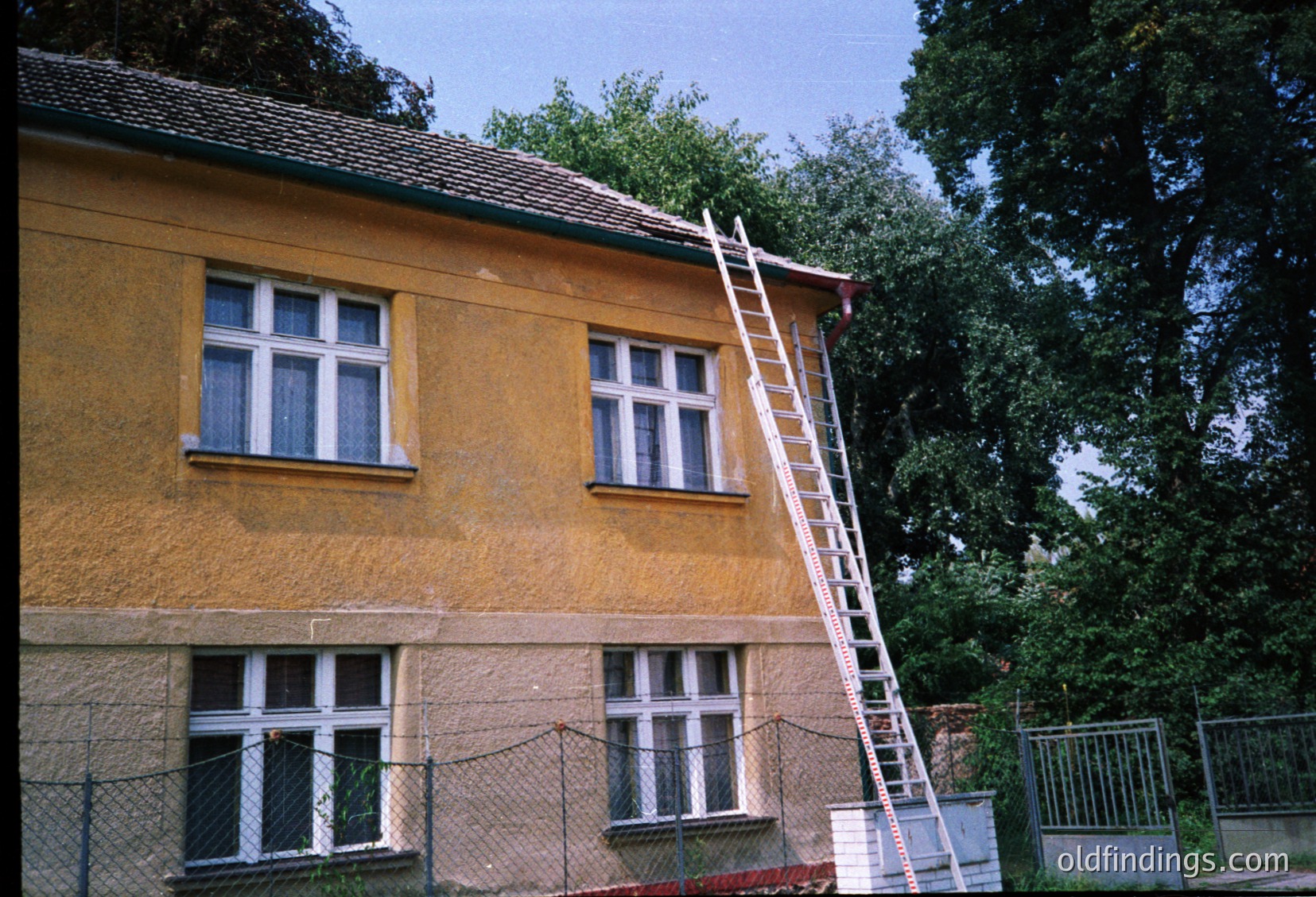 Two-story yellow stucco building with white-framed windows, likely mid-20th century. Ladder leans against roof, suggesting maintenance or renovation. Fenced perimeter with chain-link and metal gate. Lush greenery and mature trees in background.