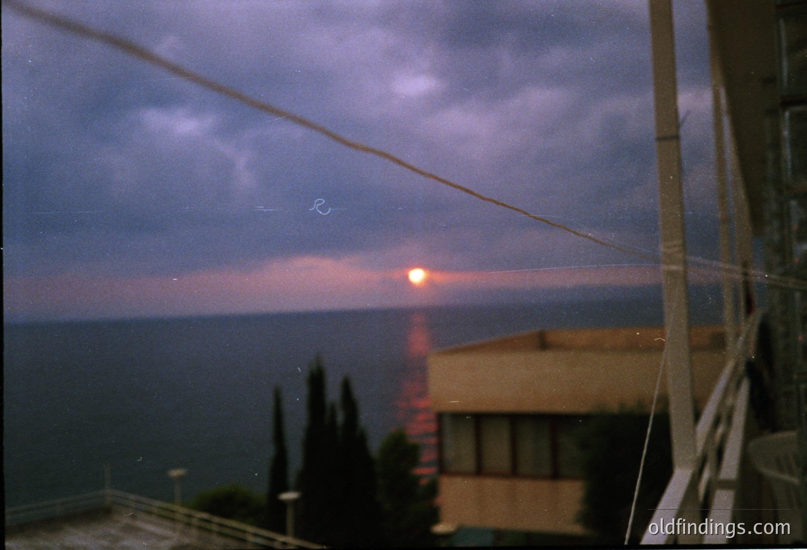 Vintage seaside sunset with lens flare over calm waters. Low-angle shot captures a lone sunbeam piercing clouds, illuminating mist. Coastal architecture—likely a seaside resort—visible in foreground. Film grain suggests 1980s–1990s era. Ideal for nostalgic or atmospheric stock imagery.