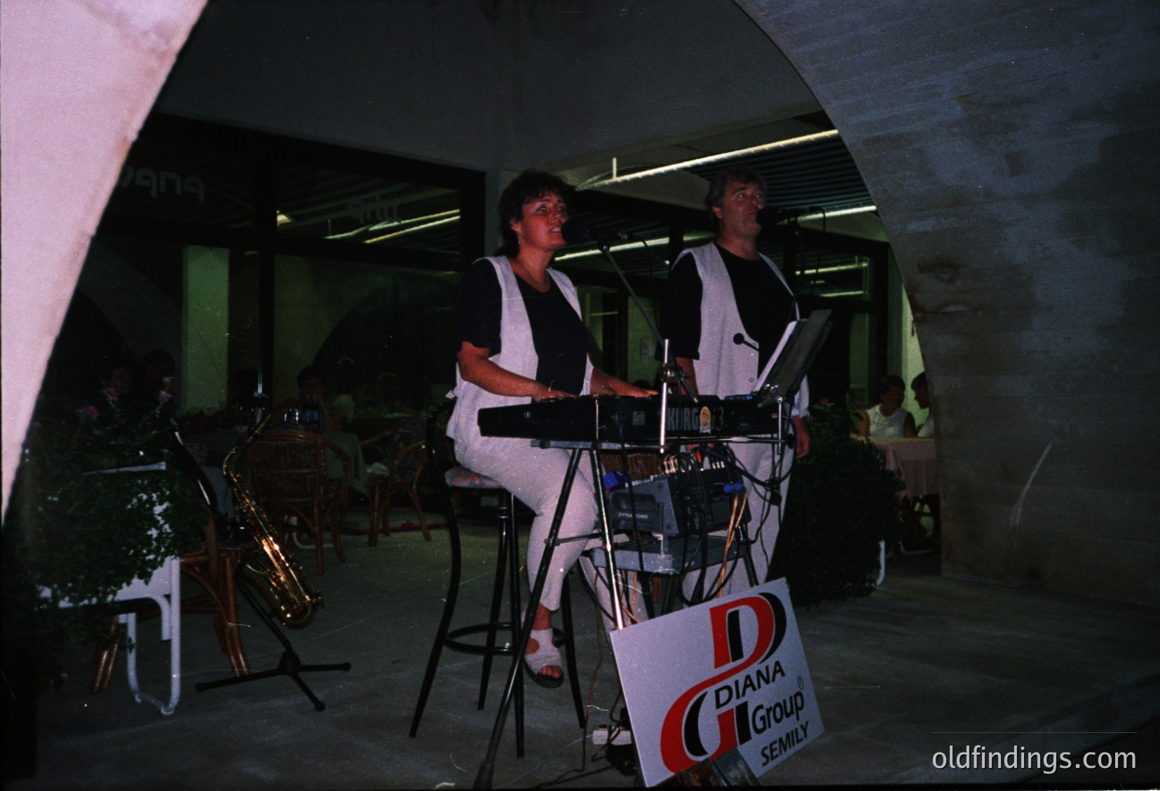 Two musicians performing live in a dimly lit, modernist indoor venue. The woman plays a keyboard, the man sings into a microphone. Signage reads "Diana Group Senily" with a red "D" logo. Brass instruments and a drum set visible in background. Mid-20th century concert setting, likely Eastern Bloc region.