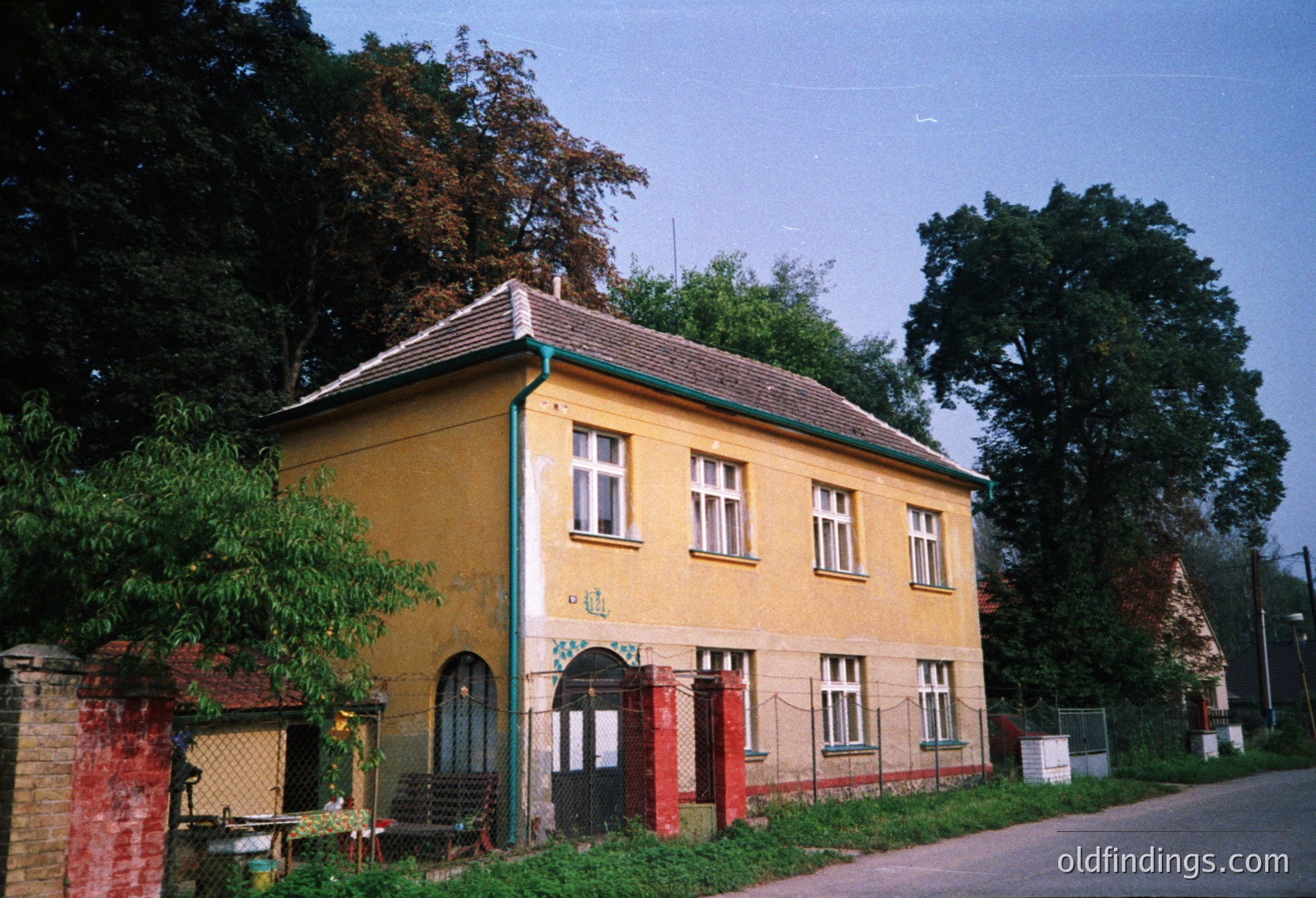 Two-story yellow building with aged paint, featuring symmetrical white-framed windows and a gabled roof. Red double doors with arched tops and a small arched window above. Surrounding greenery includes overgrown trees and ivy on a brick wall. Likely Eastern European rural architecture, mid-20th century.