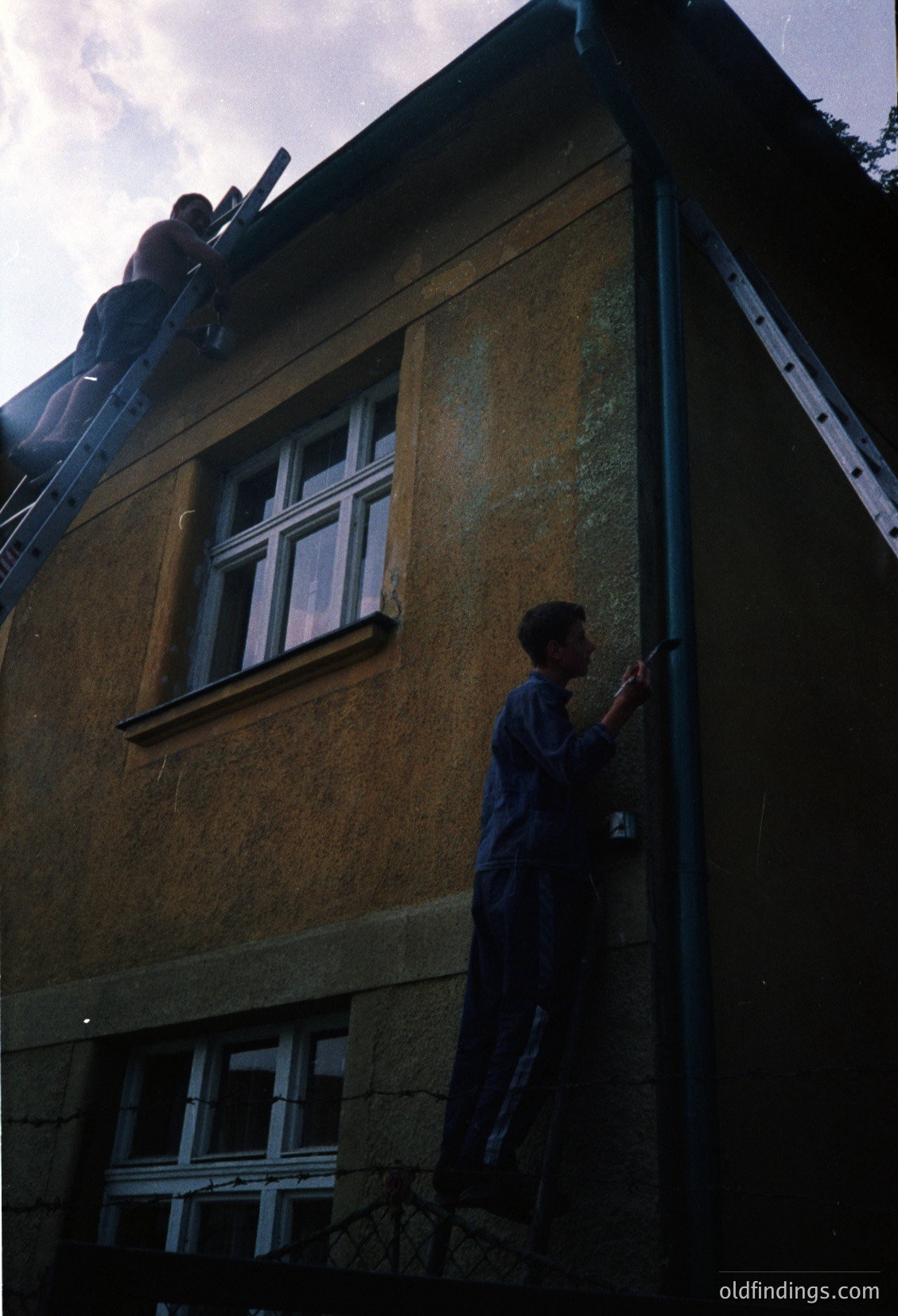 A rooftop view of a weathered concrete building facade, showing aged yellowish plaster and exposed rebar. A lone person in dark clothing climbs the exterior wall, likely performing maintenance or construction. Below, a window with white grid-patterned glass reveals a dark interior. Architectural details suggest mid-20th-century utilitarian design.
