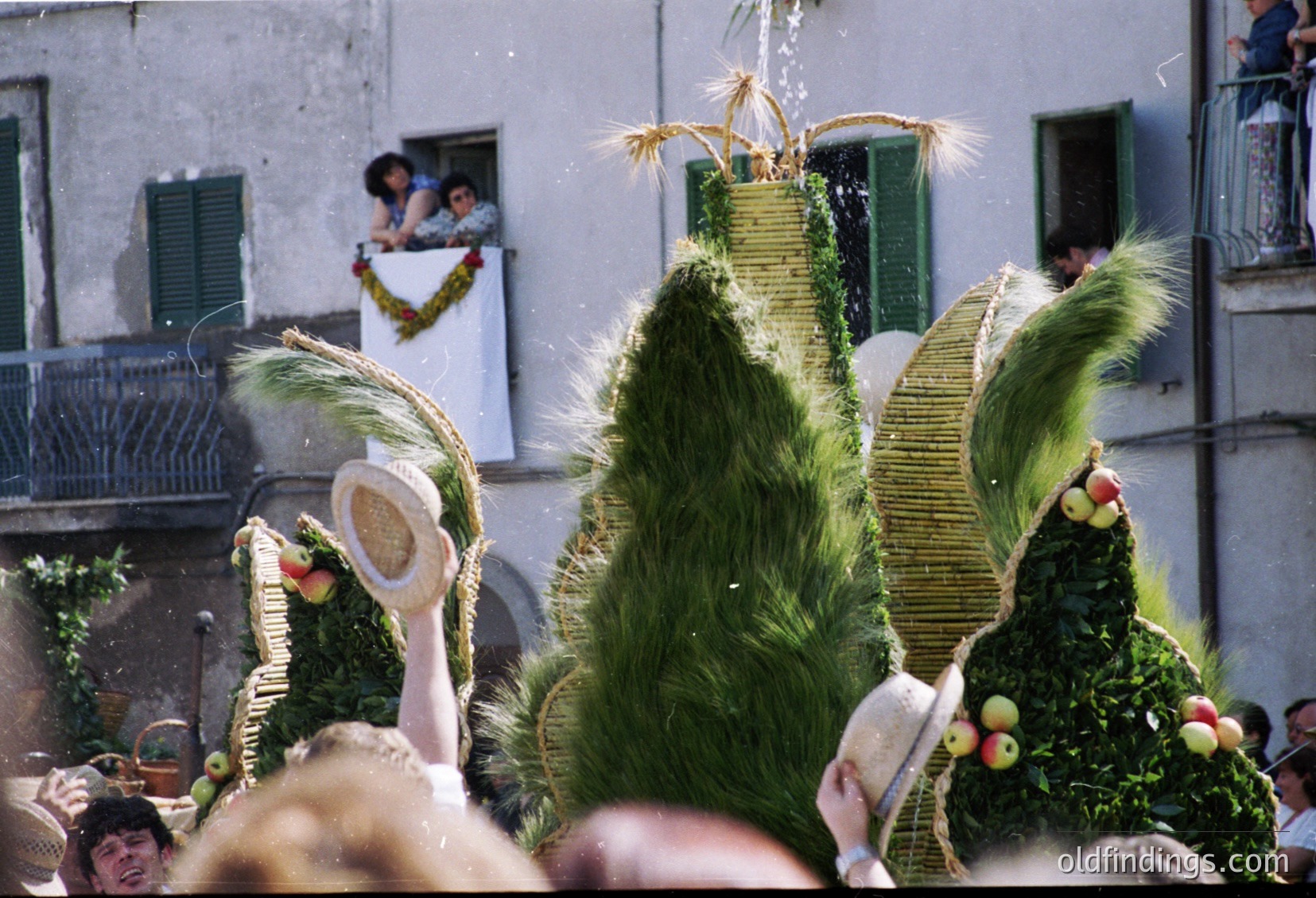 Vibrant folk festival with towering, intricately woven straw effigies adorned with greenery, fruits, and wheat bundles. Crowd gathers in a narrow European street, likely Italy’s *Festa della Sensa* or similar harvest celebration. Colorful costumes and traditional attire suggest late 20th-century rural tradition.