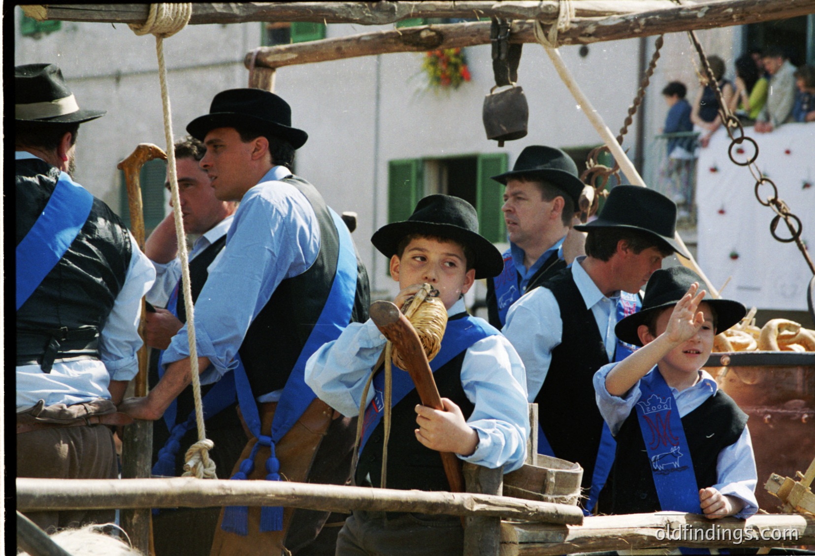 Traditional wooden boat crew in coordinated blue-and-white attire, including wide-brimmed hats, operating a hand-cranked mechanism. Likely a coastal or riverine folk festival, suggesting cultural heritage preservation.