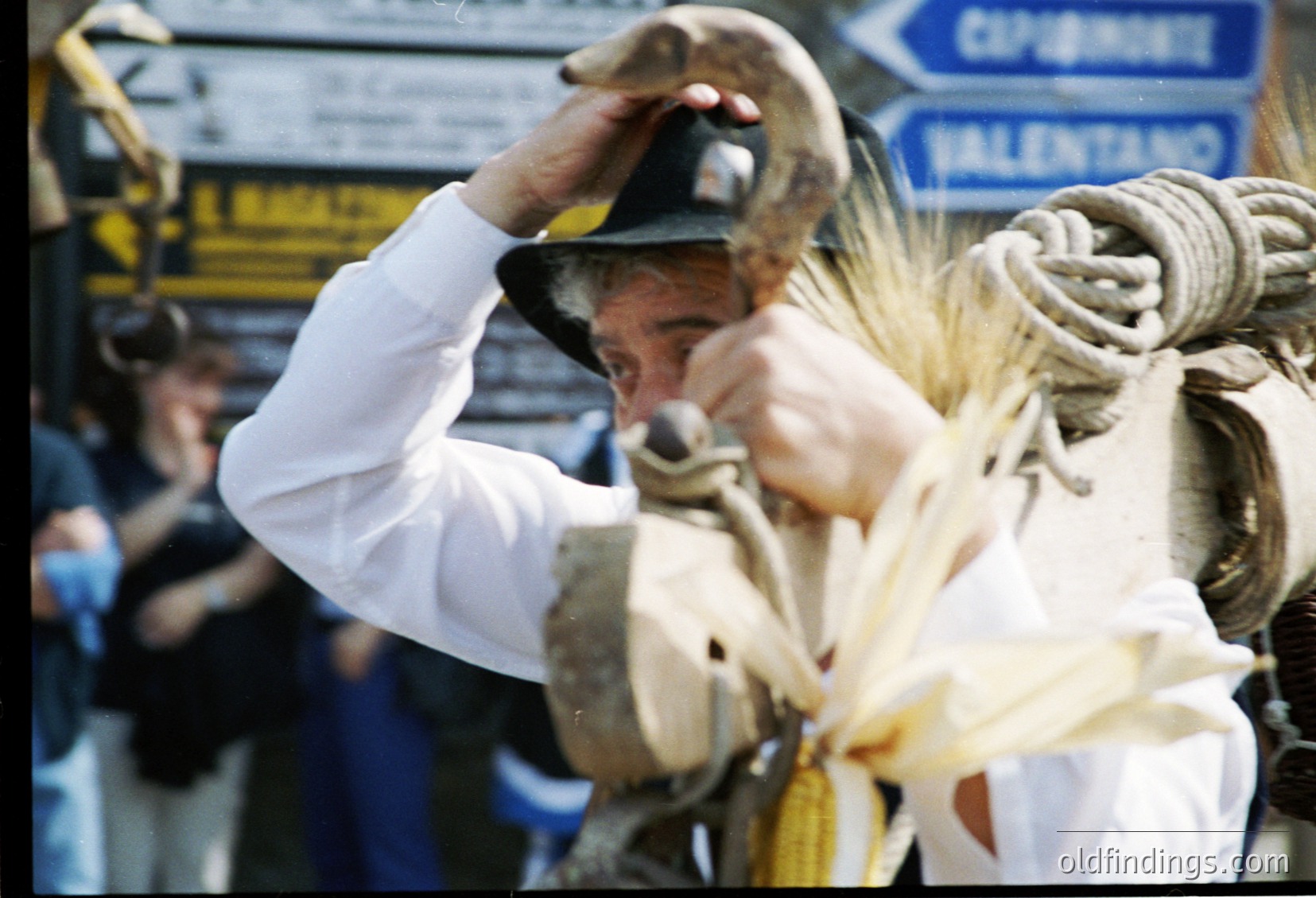 A man in traditional attire, likely a shepherd, handles a sheep in a public or market setting. His white tunic, dark hat, and rope-wrapped sheep suggest rural labor. Signs in Cyrillic indicate Eastern Europe, possibly Bulgaria. The 1960s–1980s era clothing and setting point to a mid-20th-century rural scene. [Traditional shepherd handling sheep in Eastern European market ]